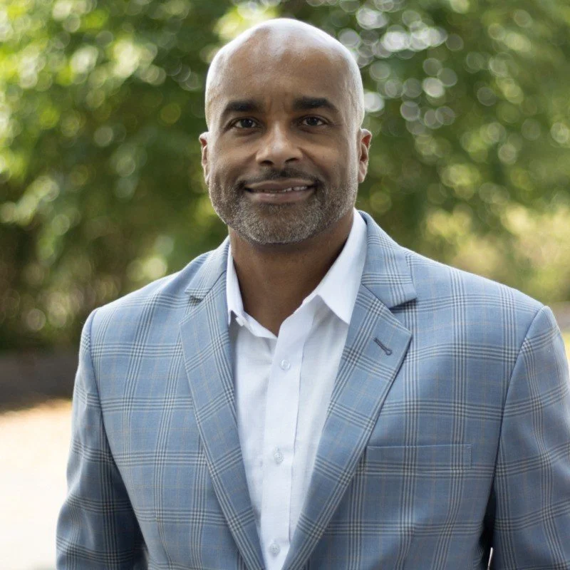 Portrait of a middle-aged man with a bald head and gray beard, wearing a light blue checked blazer and a white shirt, standing outdoors with a blurry green background.