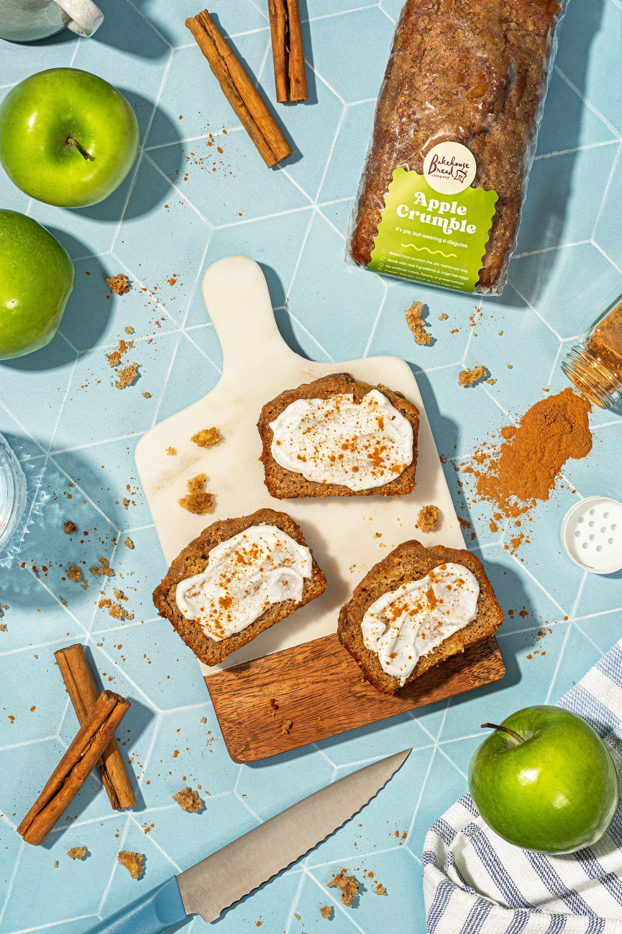 Arranged on a blue tiled surface are two green apples, a loaf of apple crumble cake with a green label, pieces of apple crumble cake with whipped topping on a white cutting board, cinnamon sticks, a serrated knife, and a cloth towel with blue stripes.