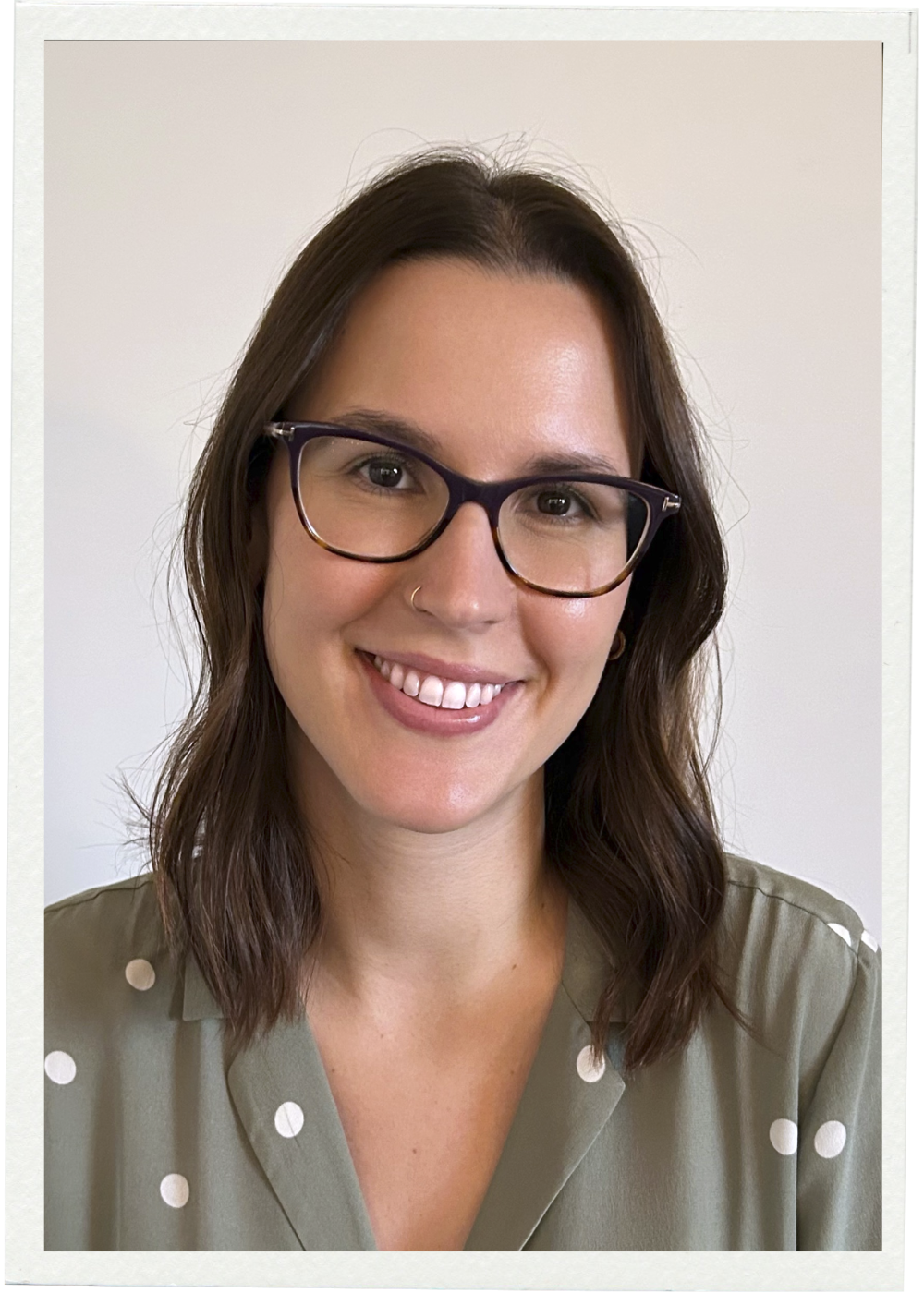 A smiling young woman with short brown hair, glasses, and a necklace, wearing a black shirt and a patterned cardigan, outdoors in front of a building.