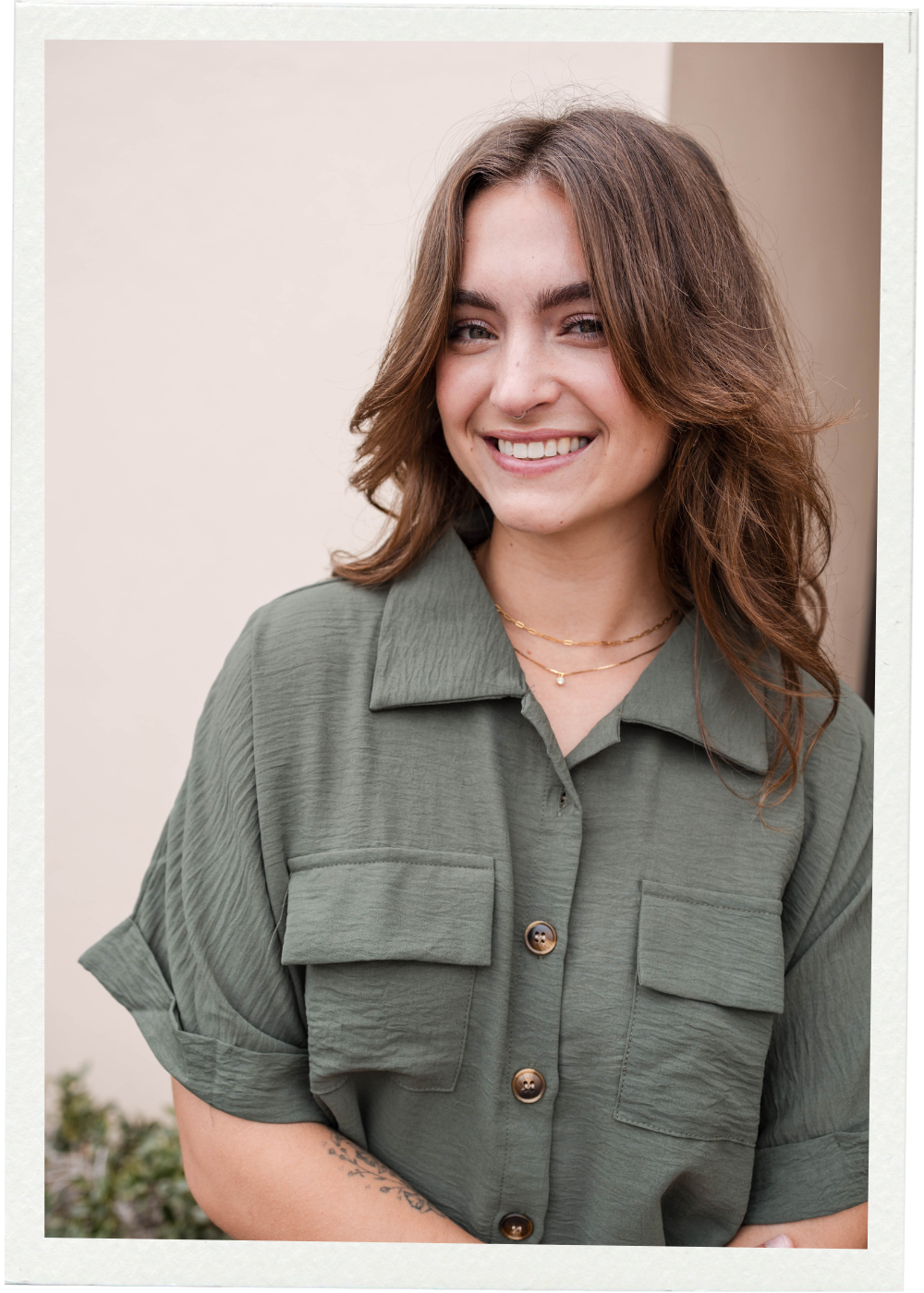 A smiling young woman with short brown hair, glasses, and a necklace, wearing a black shirt and a patterned cardigan, outdoors in front of a building.