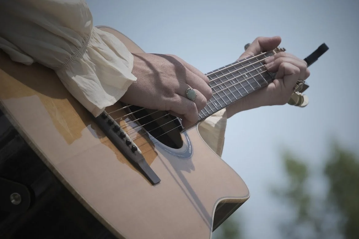 Person playing an acoustic guitar outdoors, wearing a cream-colored blouse, with a ring on their finger.