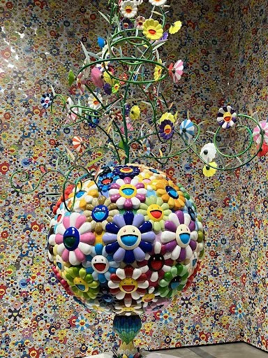 A vibrant, colorful sculpture featuring a spherical base covered with various smiling, flower-shaped faces, topped with a whimsical metal tree with painted, flower-like blossoms and loops against a multicolored mosaic-patterned background.