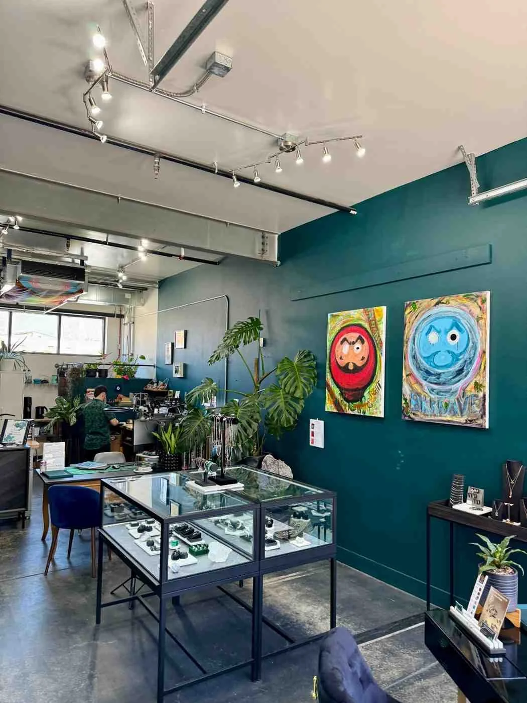 Modern jewelry store interior with industrial ceiling, dark green accent wall, glass display cases containing jewelry, colorful artwork, plants, and track lighting throughout the space
