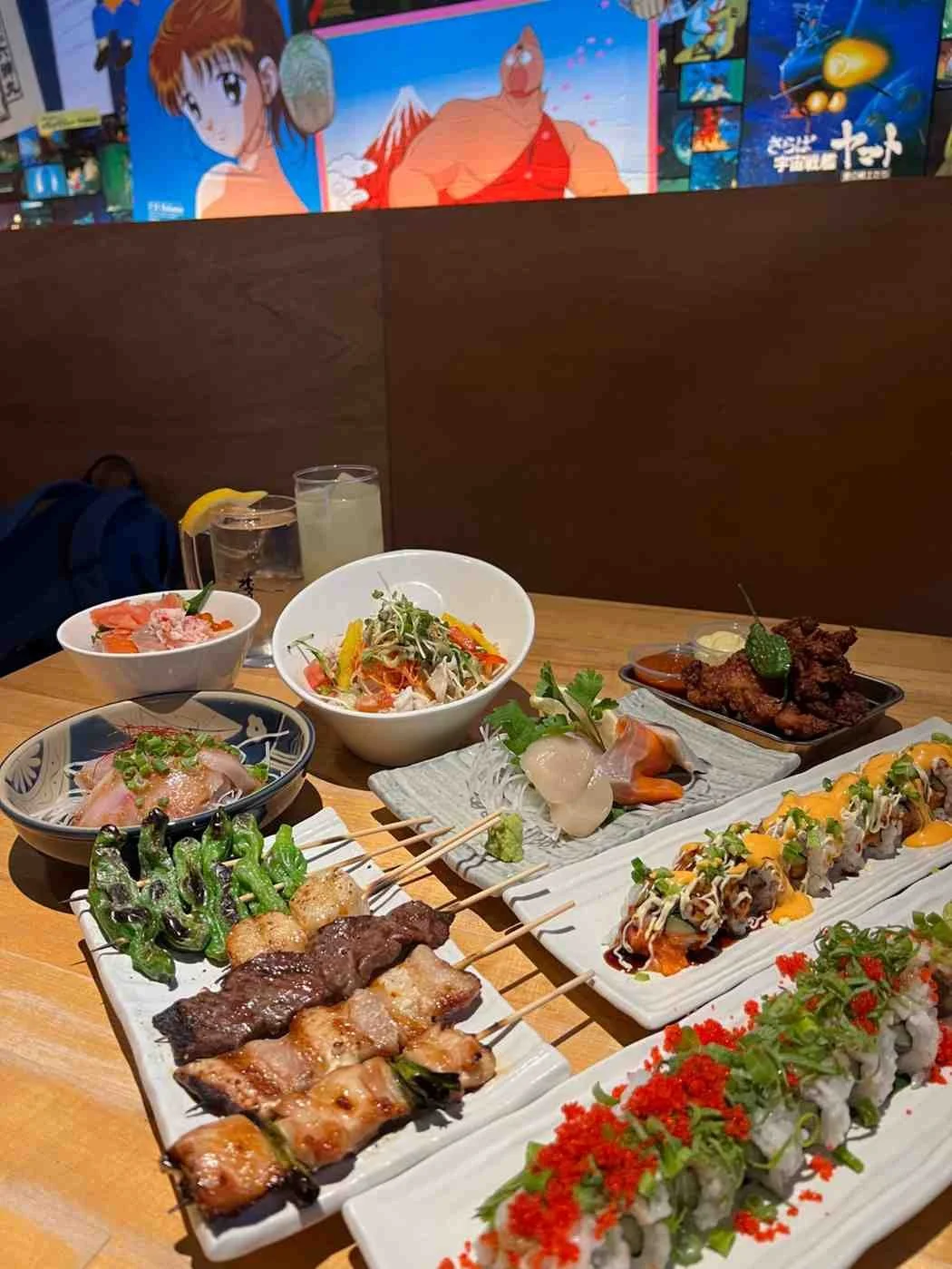 Japanese restaurant table with sushi, yakitori skewers, sashimi bowls and drinks under anime wall screens