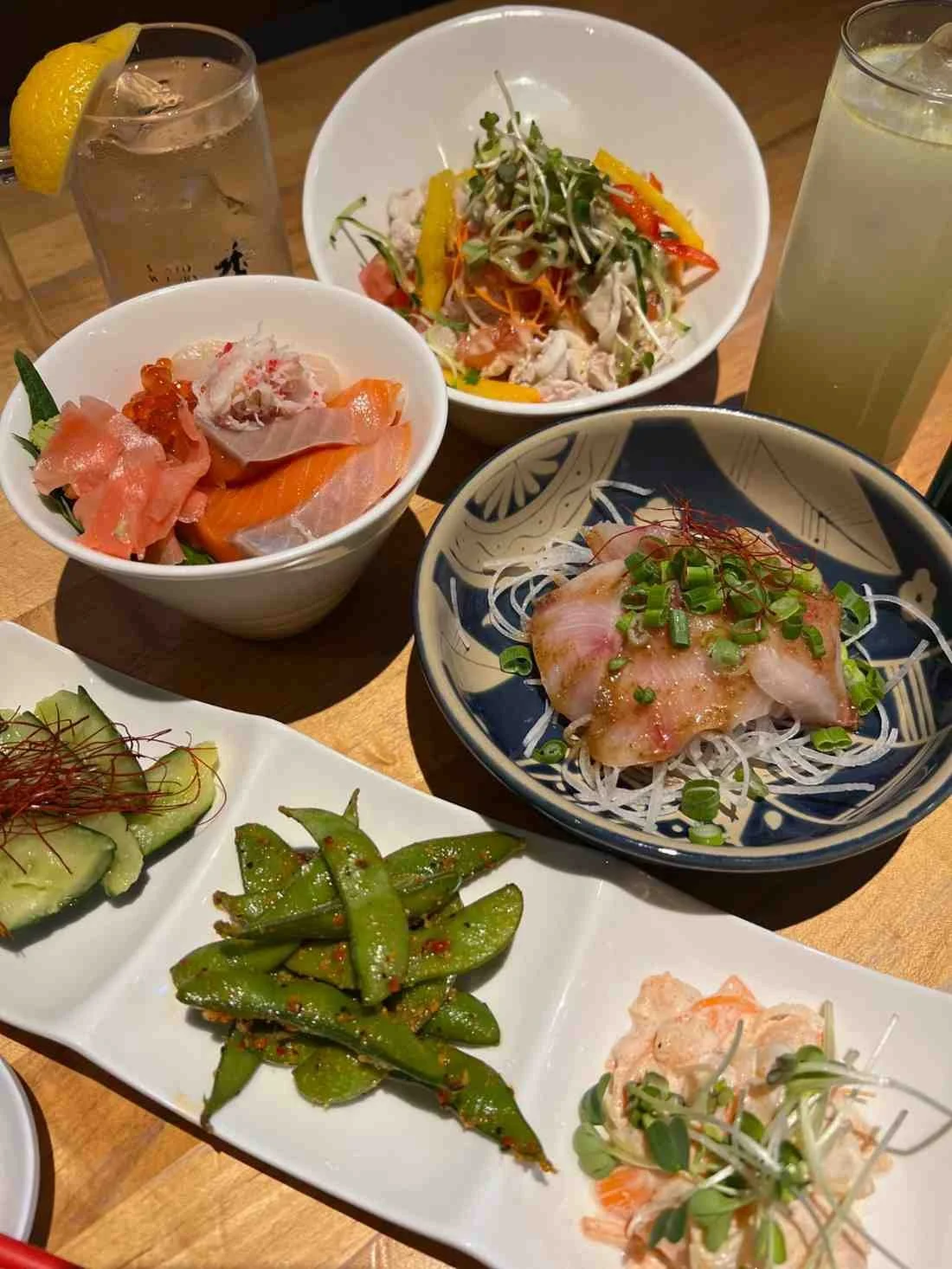 Japanese meal spread with sashimi bowls, edamame, cucumber salad, and drinks on wooden table