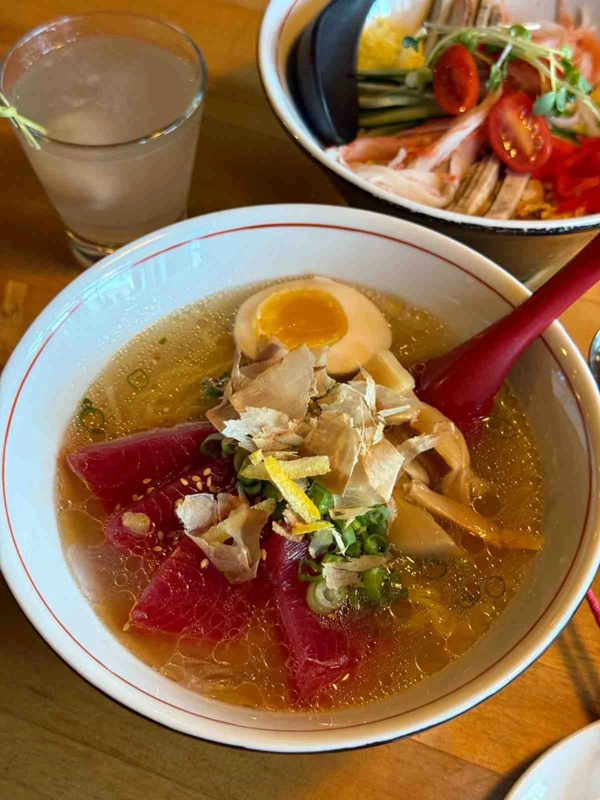 Close-up of ramen bowl with soft-boiled egg, sliced meat, and clear broth with chopsticks and drink visible