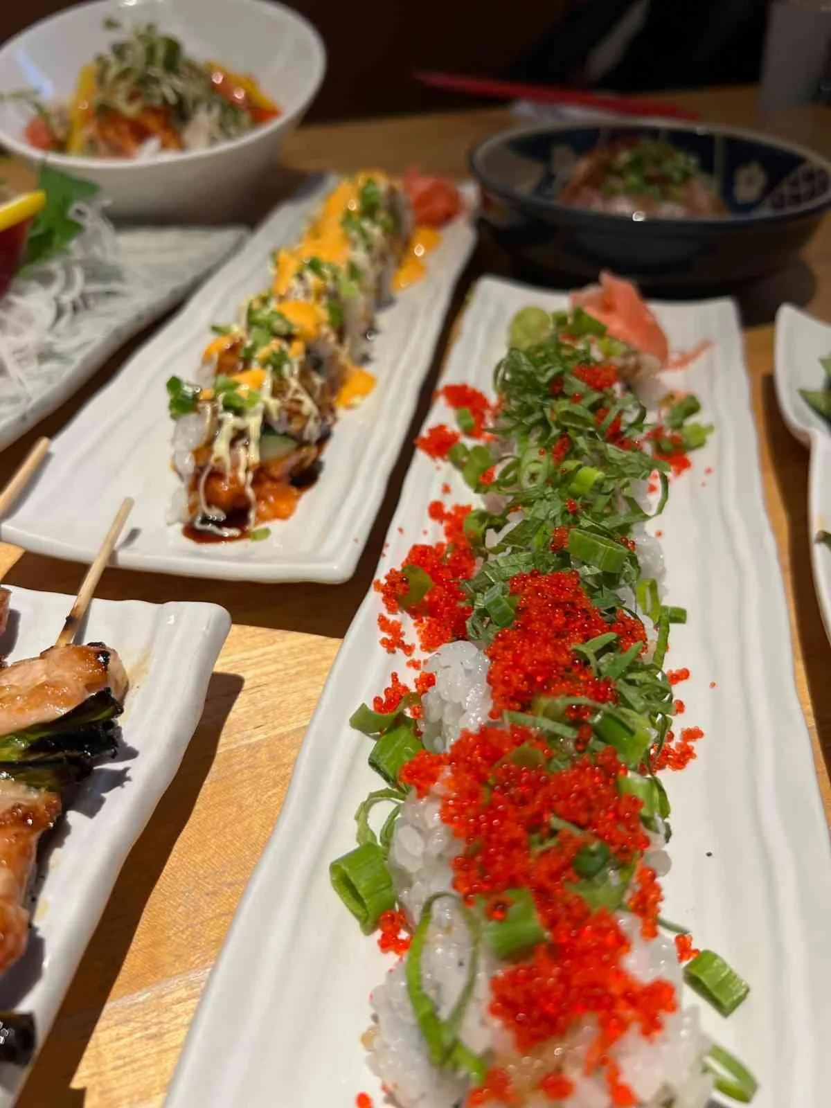 Assorted Japanese dishes on white rectangular plates including sushi rolls topped with orange roe and green garnish, alongside soup bowls and grilled items