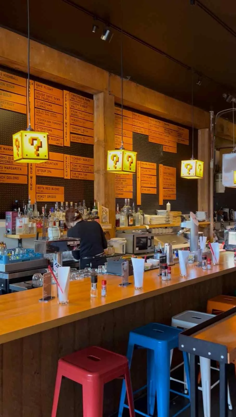 Interior of a vibrant restaurant with orange-lit menu boards, Super Mario question block pendant lights, colorful metal bar stools, and a bartender working behind the counter