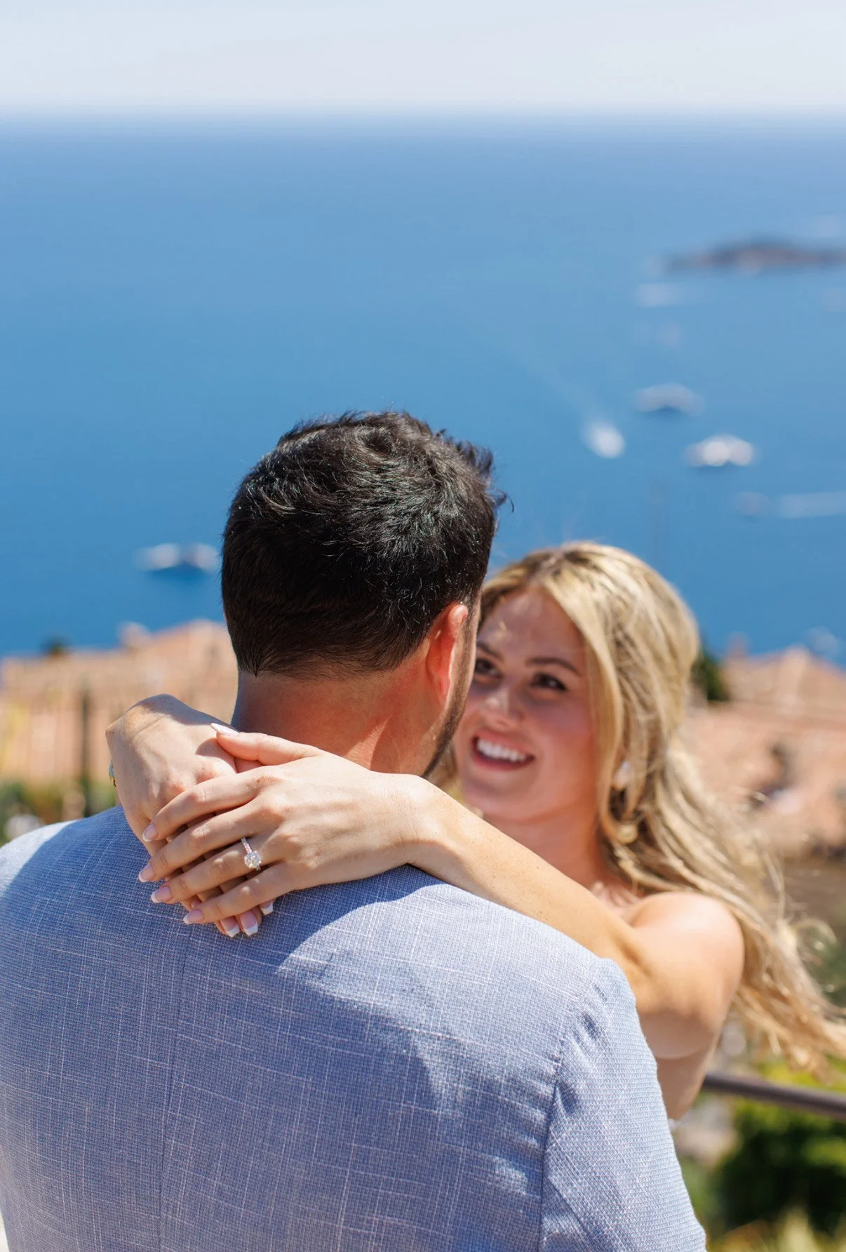 A smiling woman with blonde hair embraces a man with dark hair outdoors, overlooking a blue sea with boats and a distant coastline.