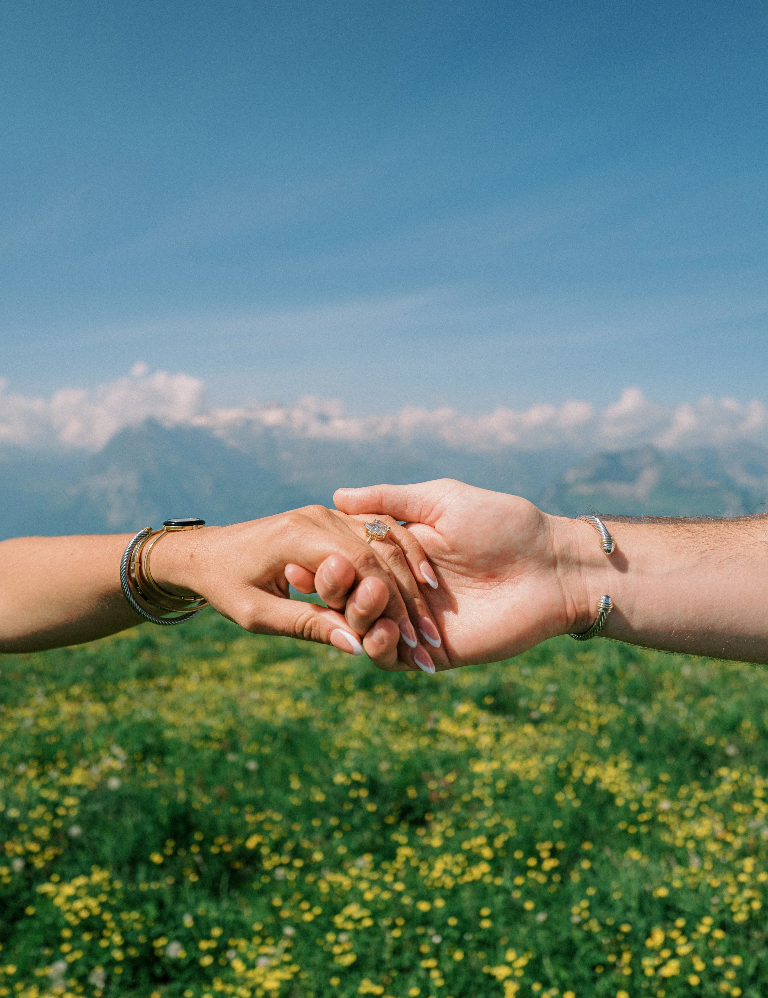 Two people holding hands against a scenic mountain and cloudy sky background, with both wearing jewelry.