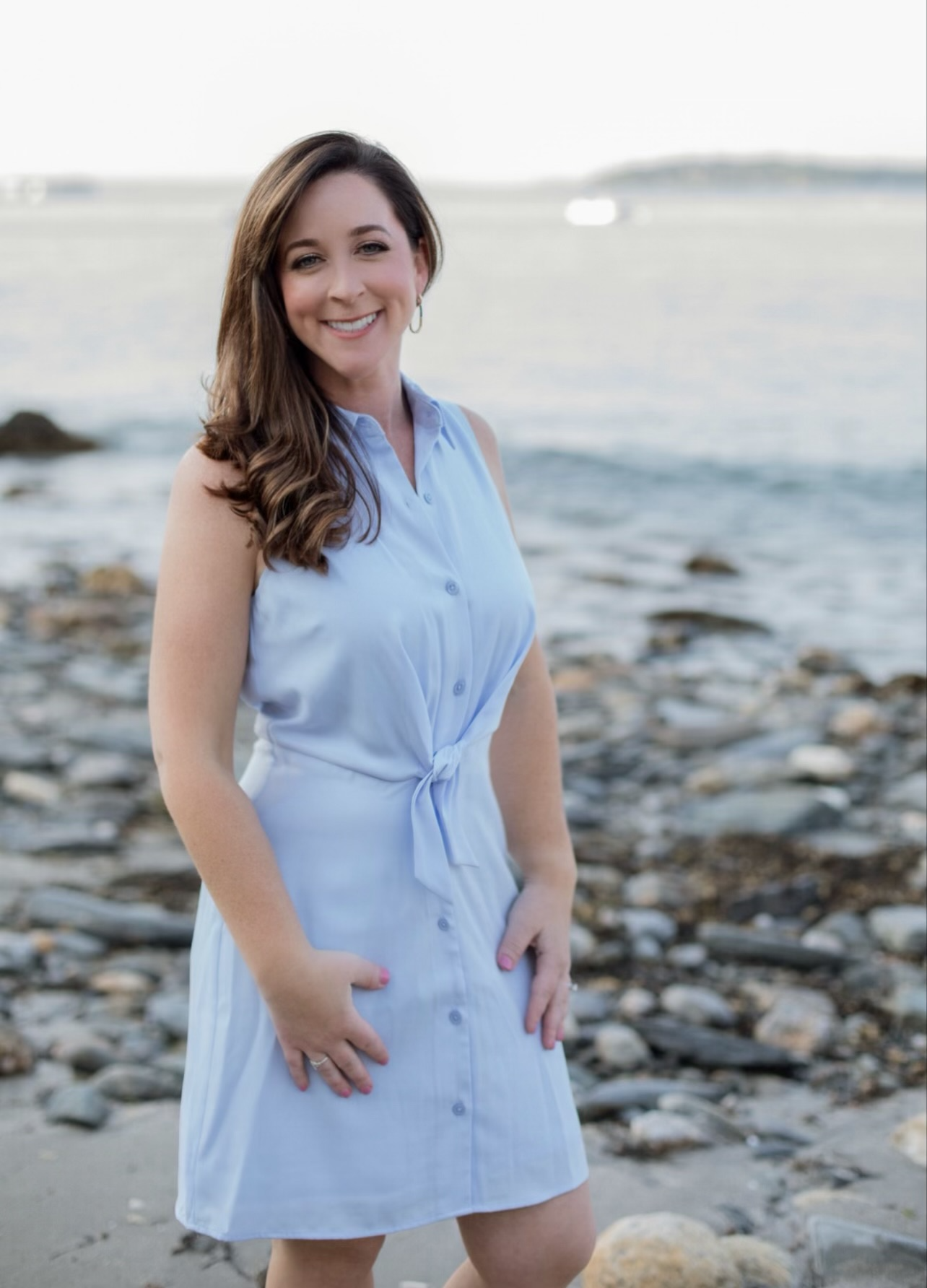 A woman with long brown hair, wearing a sleeveless light blue dress, smiling at the camera on a rocky beach with water and a distant landmass in the background.