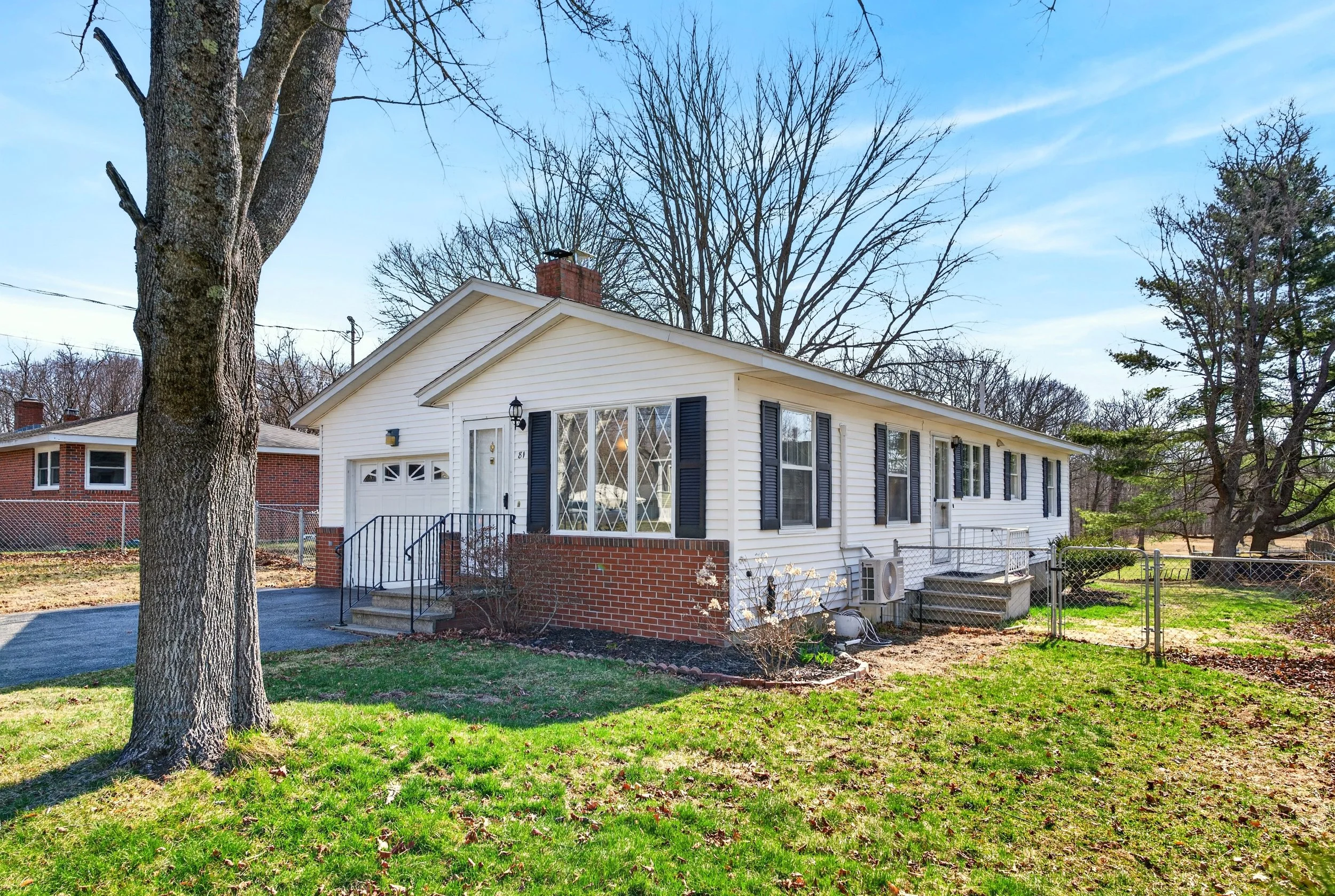 A house with white siding, black shutters, and a brick foundation is surrounded by a yard with trees, some without leaves, and a partly cloudy sky.