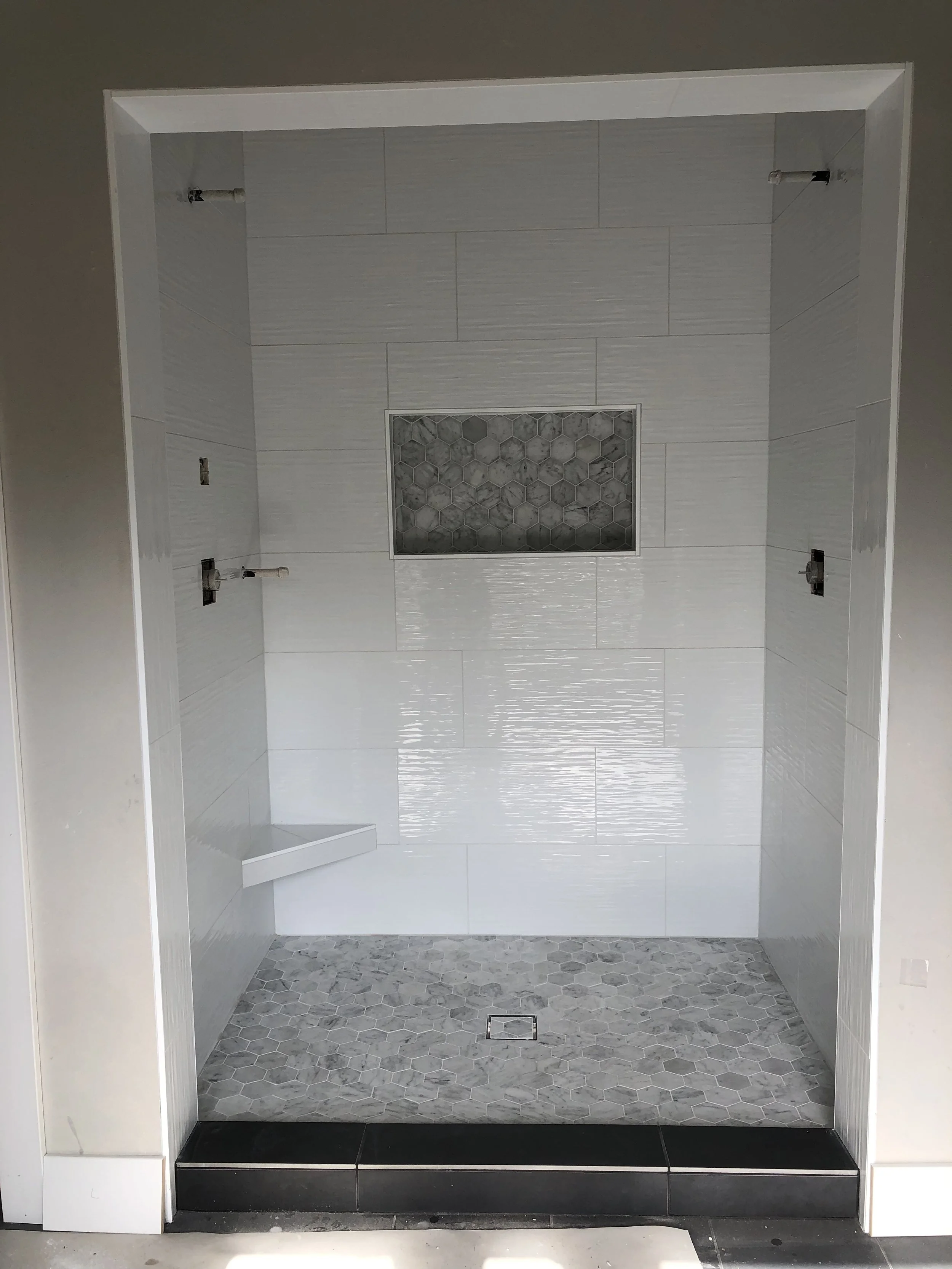 Empty walk-in shower with white tile walls, a small corner shelf, gray hexagonal floor tiles, a central drain, and a tiled niche with gray hexagonal tiles.