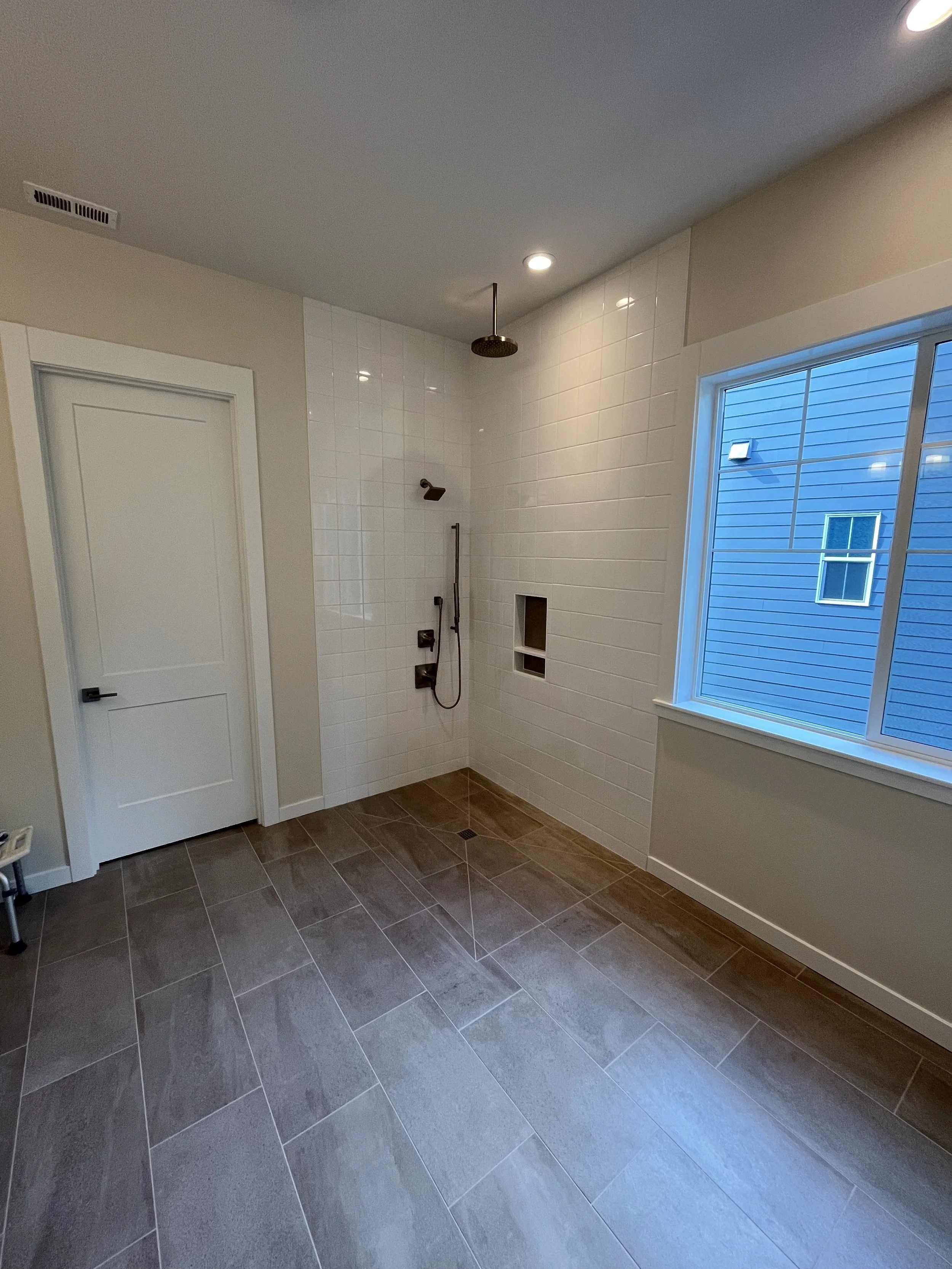 Bathroom with large gray tiled floor, white tiled shower wall with black fixtures, a window showing an outside blue house, and a ceiling with recessed lights and a rain shower head.