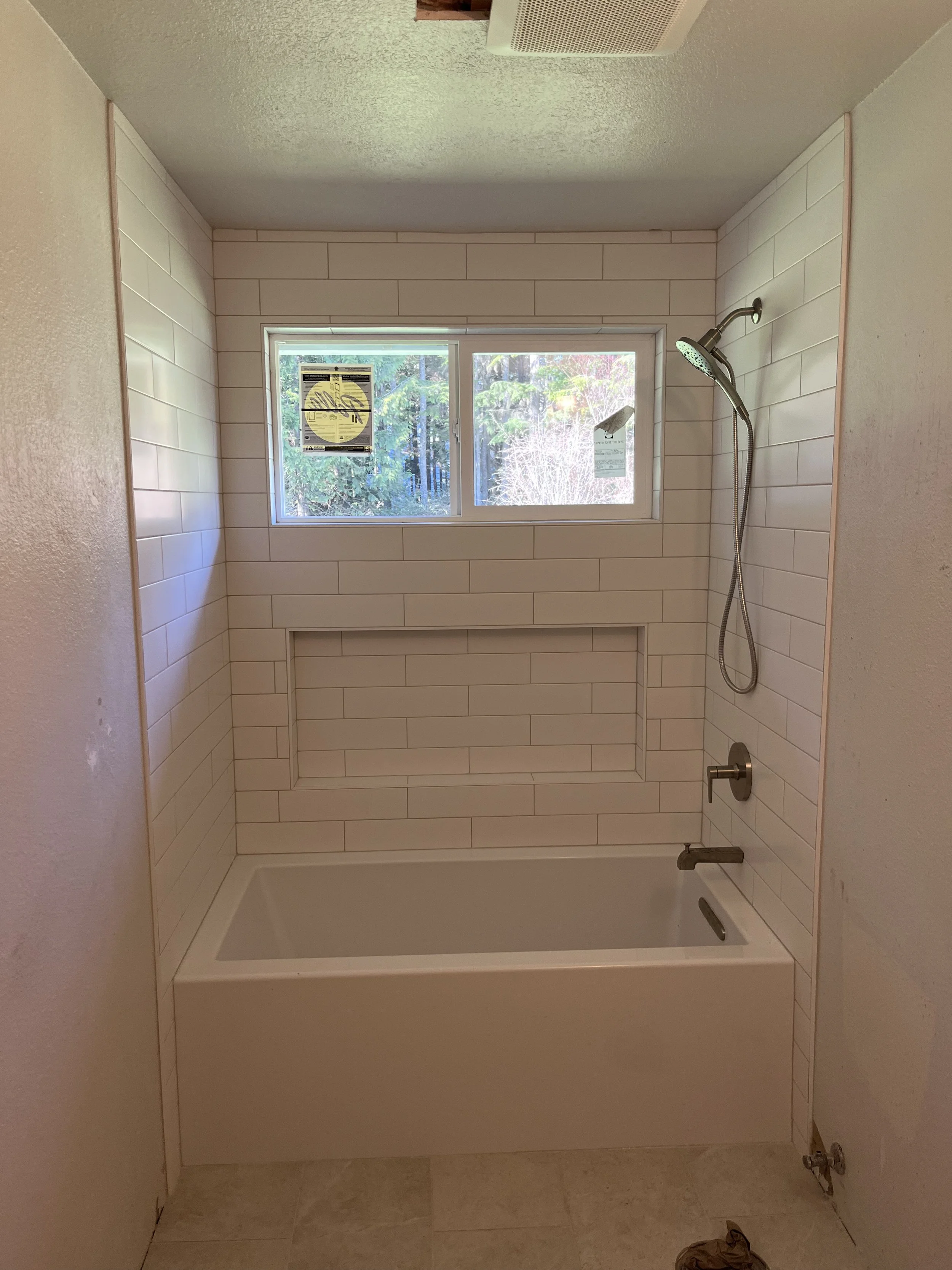 Bathroom with white tiled walls around a bathtub, a window above the tub showing outdoor trees, a showerhead and a faucet on the right, with a small bag or cloth on the floor near the bottom right corner.