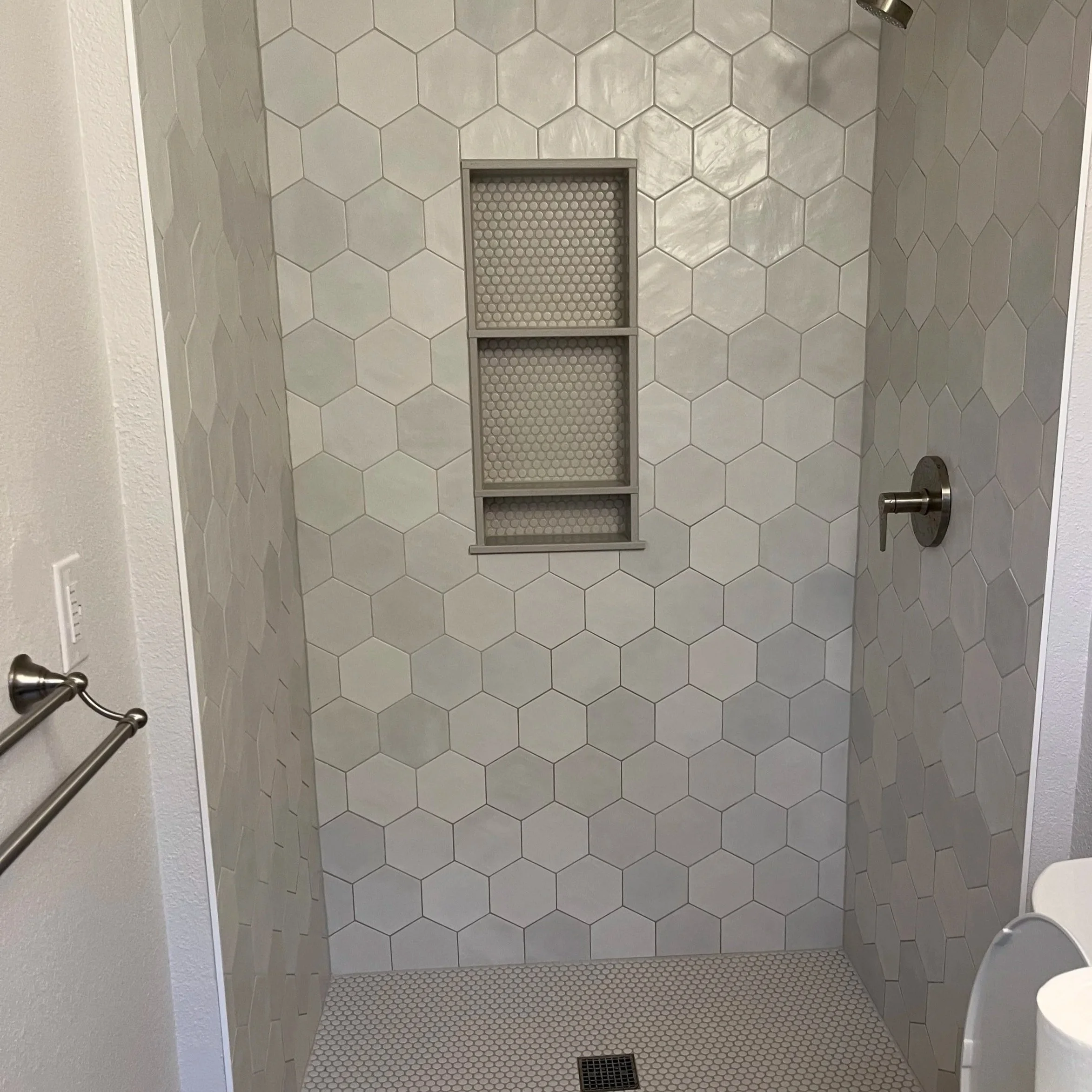A tiled shower area with three built-in shelves and a drain on the floor. The shower walls are covered with large, light-colored hexagonal tiles.