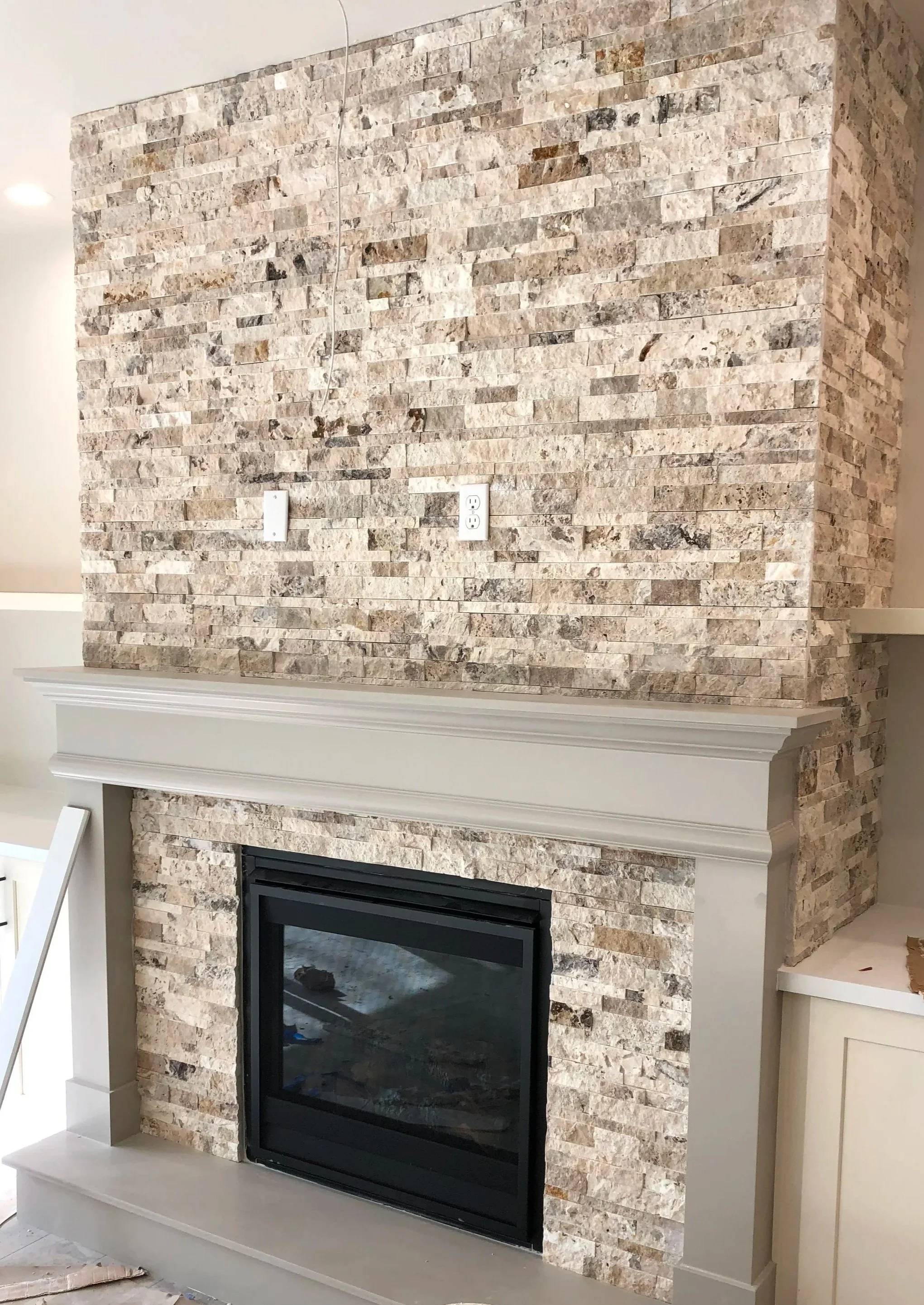 Living room fireplace with a stone face and a white mantel, with electrical outlets above on the stone wall.