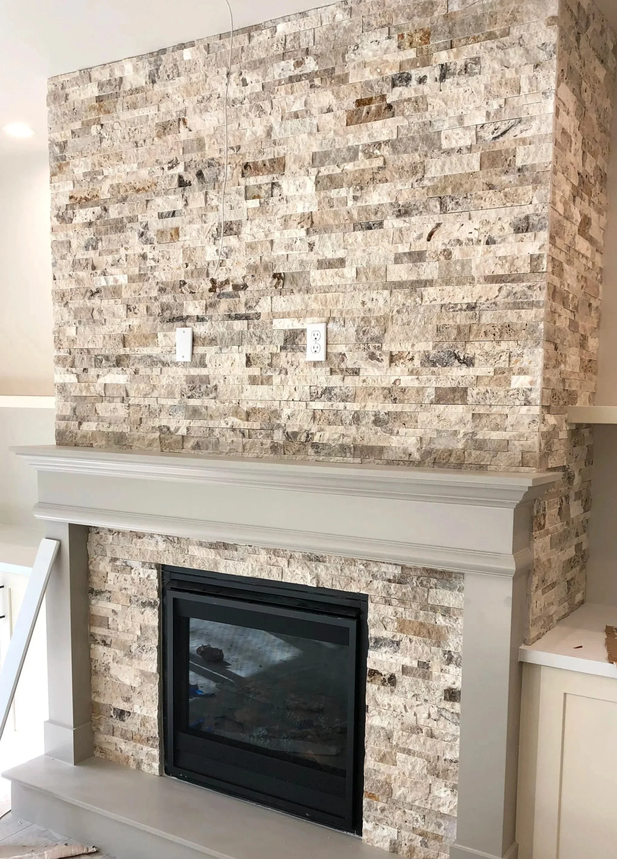 Interior view of a living room fireplace with beige stacked stone surround, white mantel, and black framed glass opening.