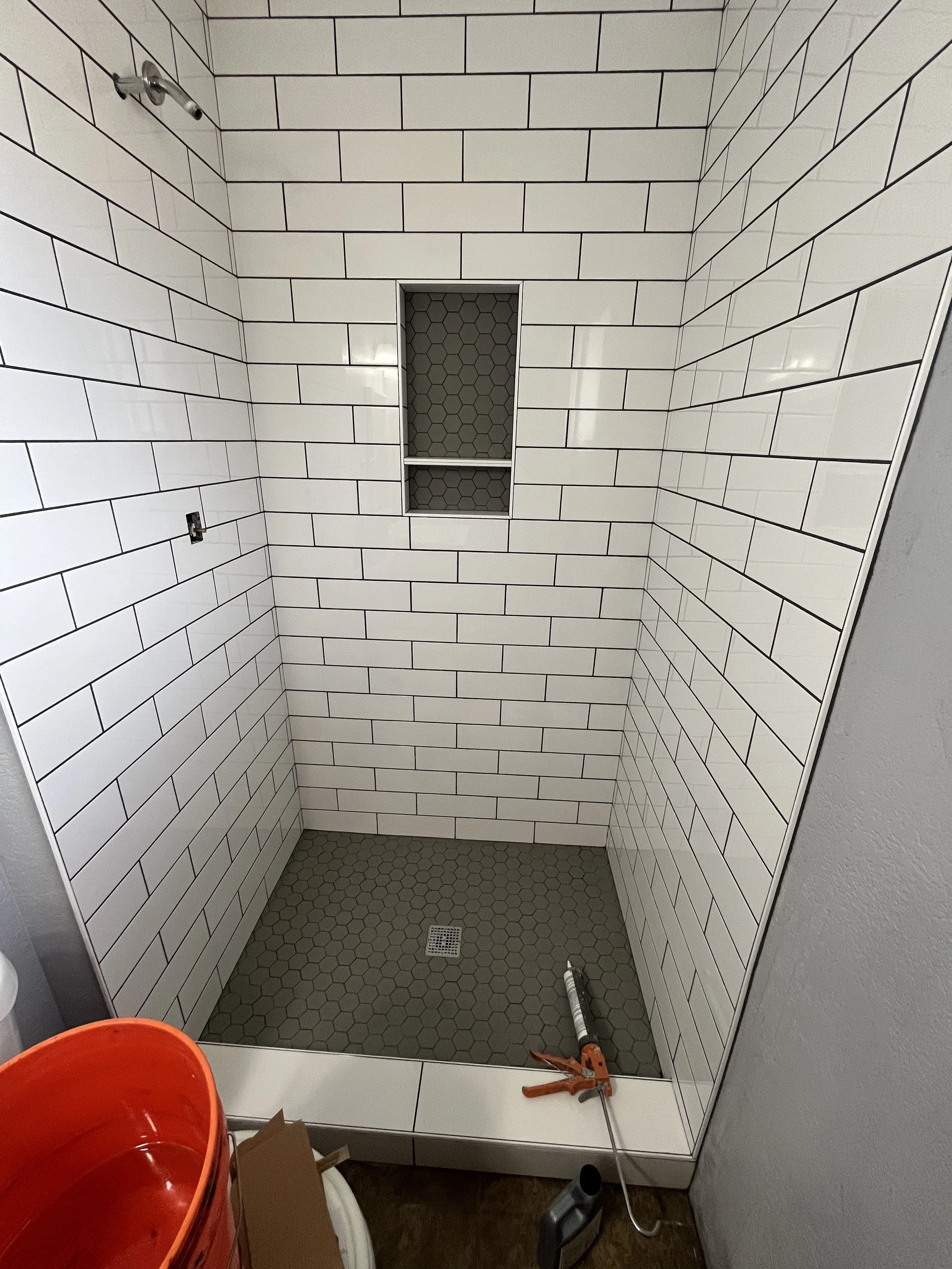 A shower stall with white subway tiles, a built-in shelf with honeycomb pattern tiles, and a square drain on a hexagon tile floor. Tools and a red bucket are in front of the stall.