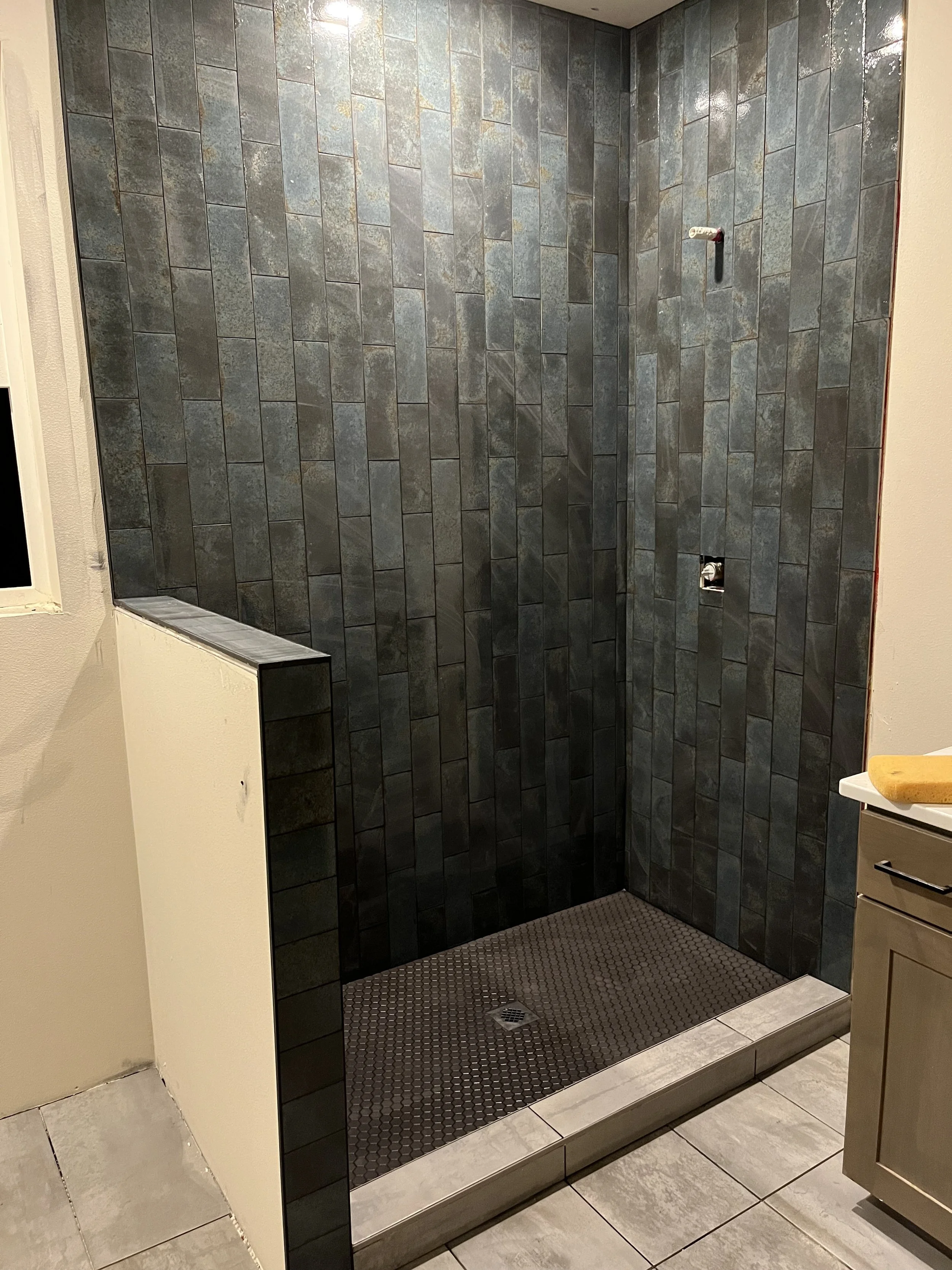 A walk-in shower with dark gray tiled walls, black textured floor, and a small window in the top left corner. There is an empty wall-mounted soap holder and a drain in the floor. Part of a beige cabinet or vanity with a yellow sponge on top is visibl