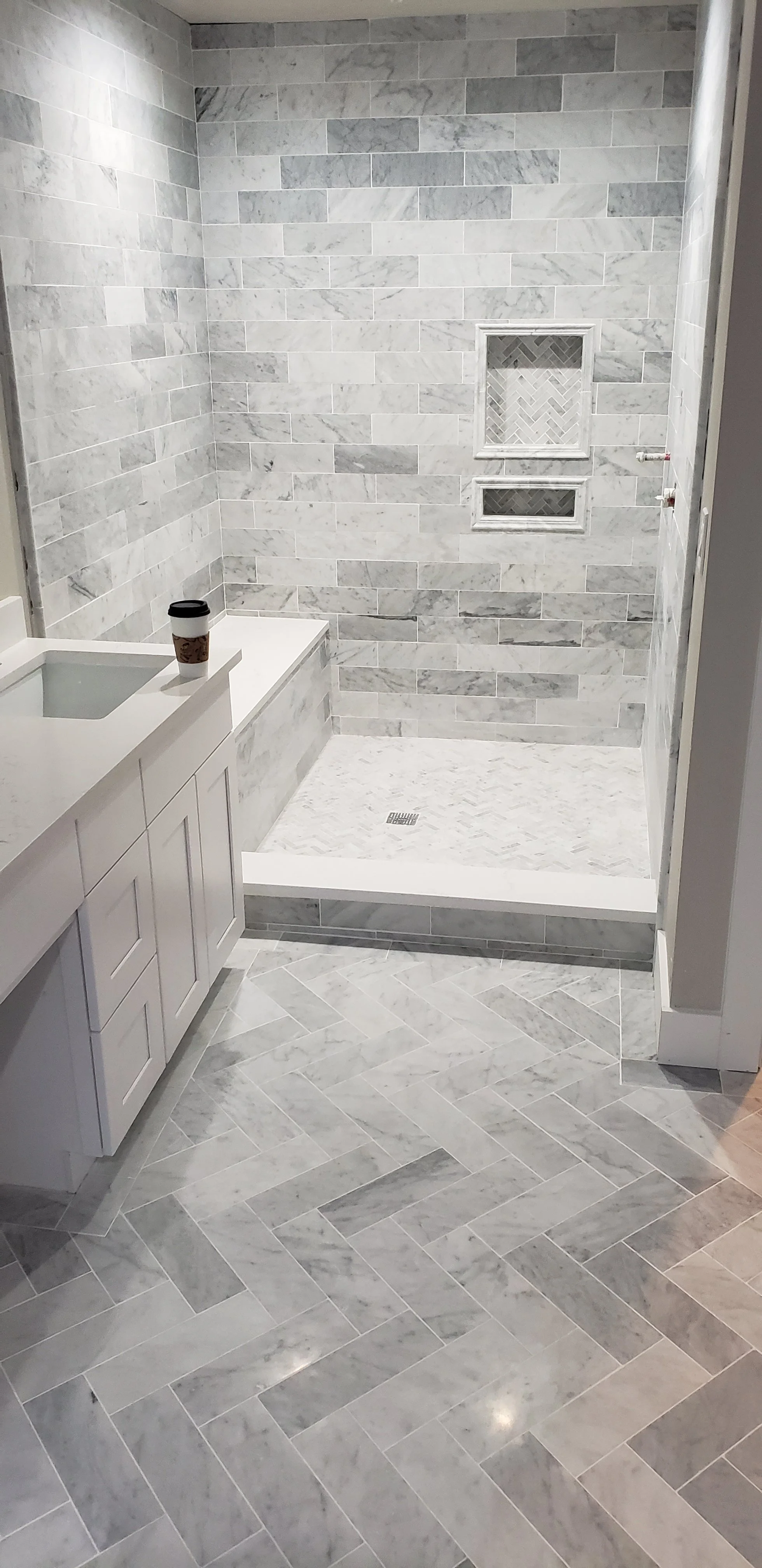 A bathroom with a walk-in shower featuring gray marble tiles, a built-in niche for toiletries, and a drain in the shower floor. The bathroom floor and shower walls are tiled with marble, and there is a white vanity with a small cup on top.