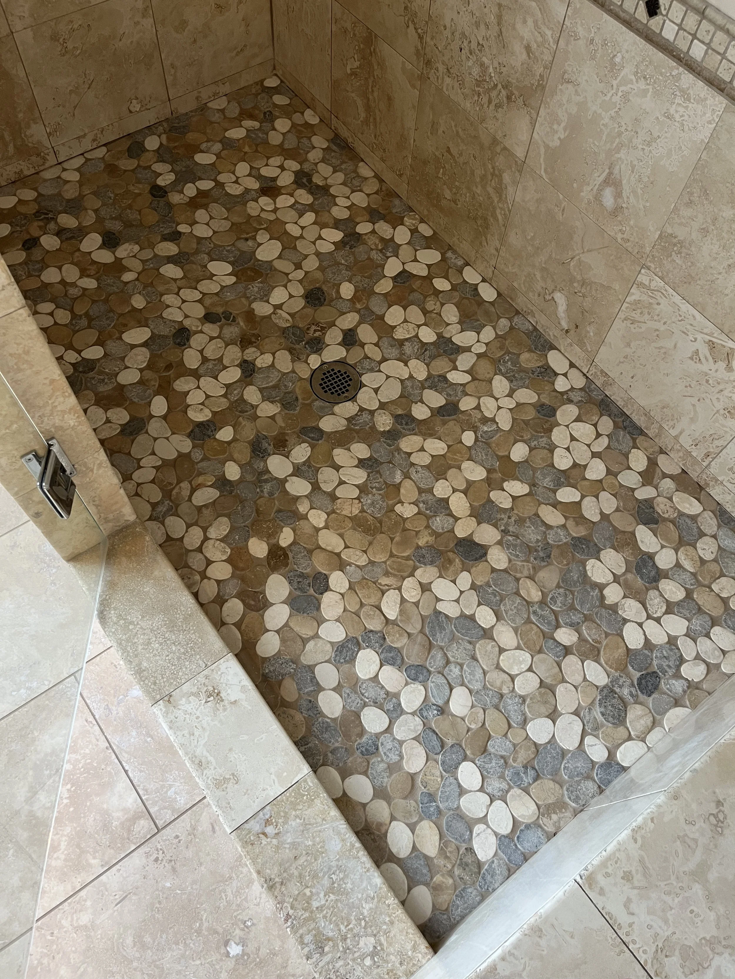 A shower stall with a floor made of rounded pebble stones in shades of beige, brown, and gray. The walls are tiled with large beige marble tiles, and there is a round drain in the center of the floor.