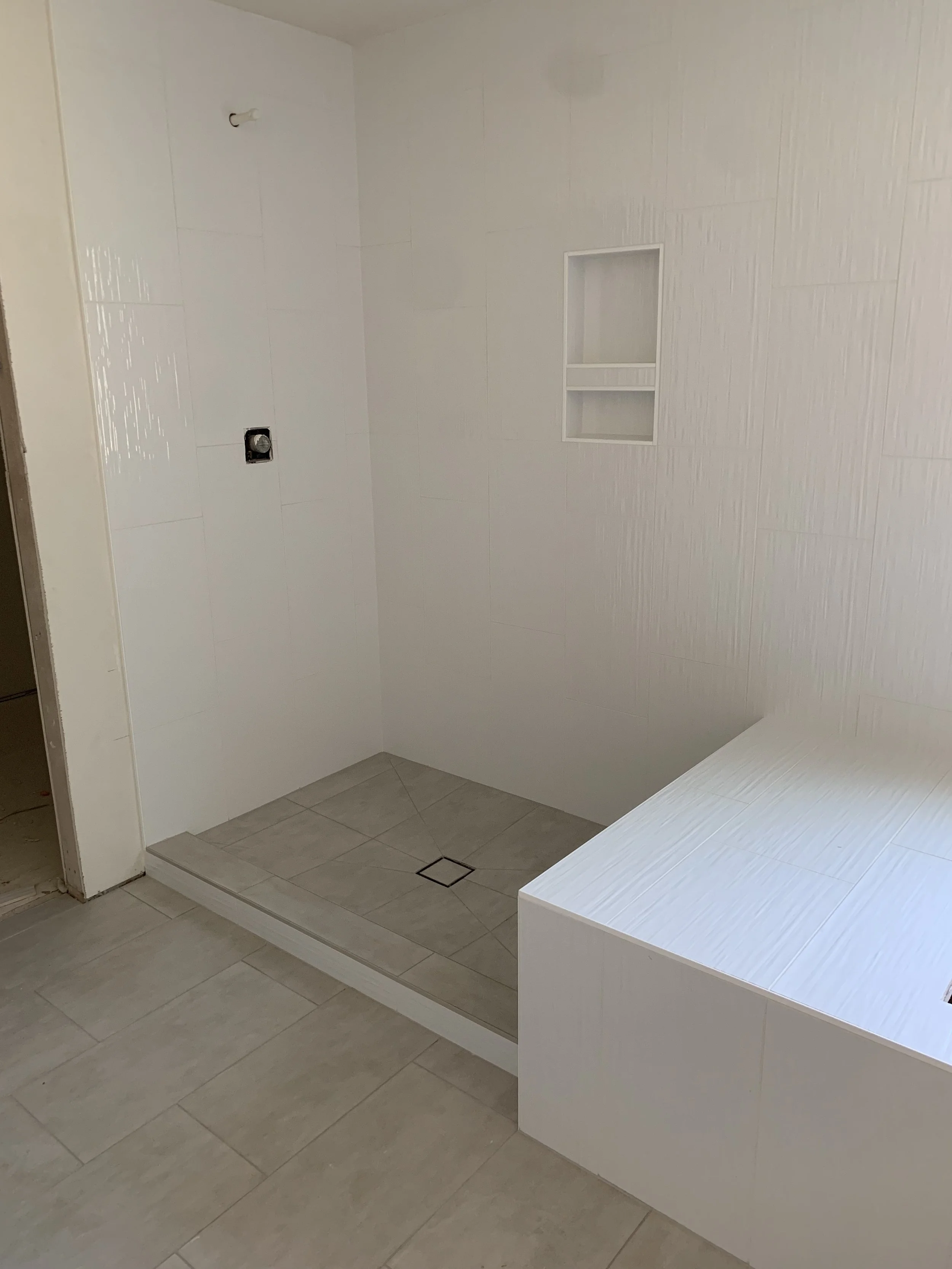 A modern bathroom with a walk-in shower that has a small built-in shelf, a drain on the floor, and a wall outlet near the ceiling, all featuring light-colored tiles.