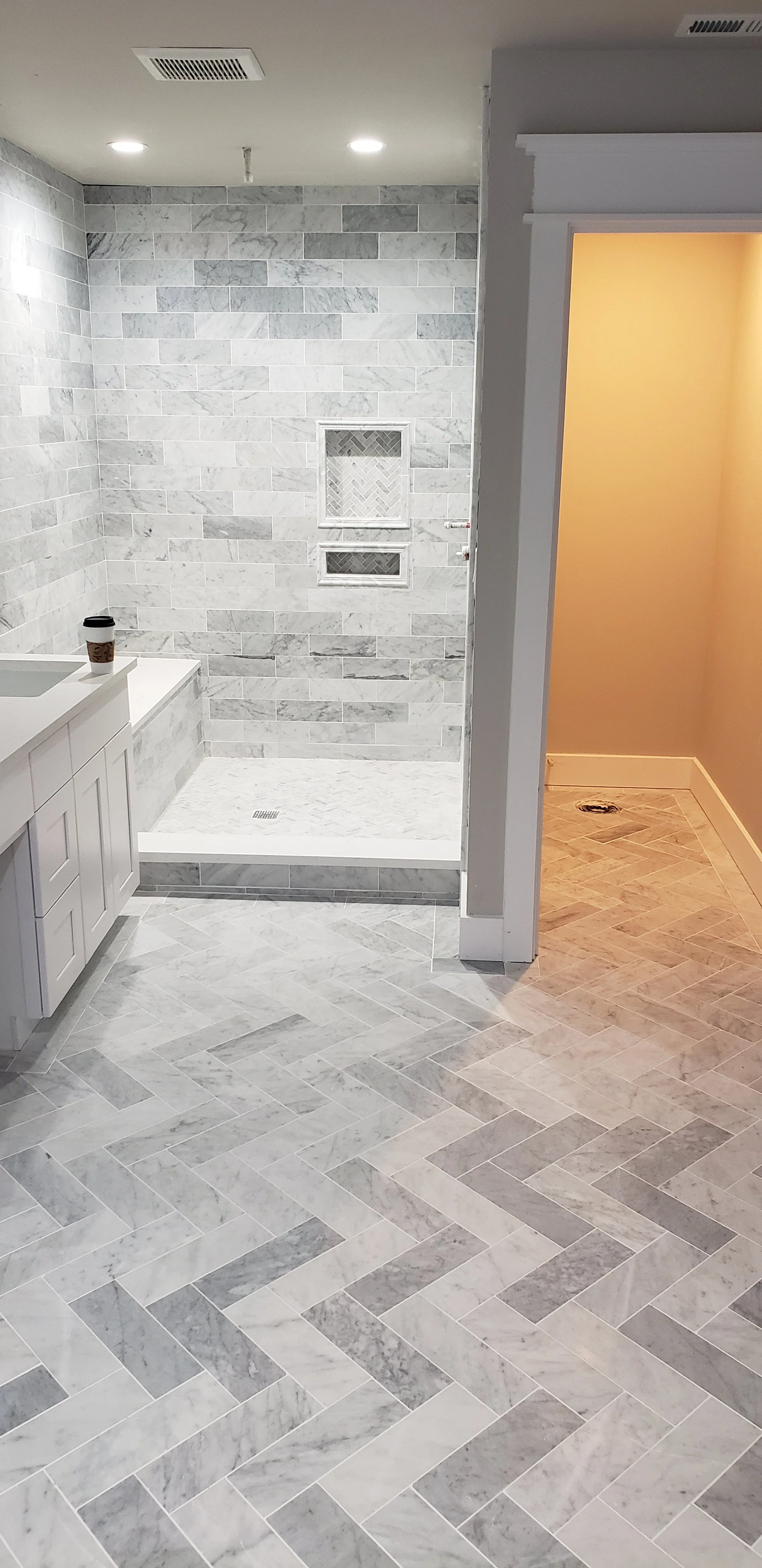 Bathroom with gray marble tiles on the walls and floor, featuring a walk-in shower area with a white marble tile base and two built-in shower niches. To the left, there is a white vanity with a small coffee cup on top. To the right, an open doorway r