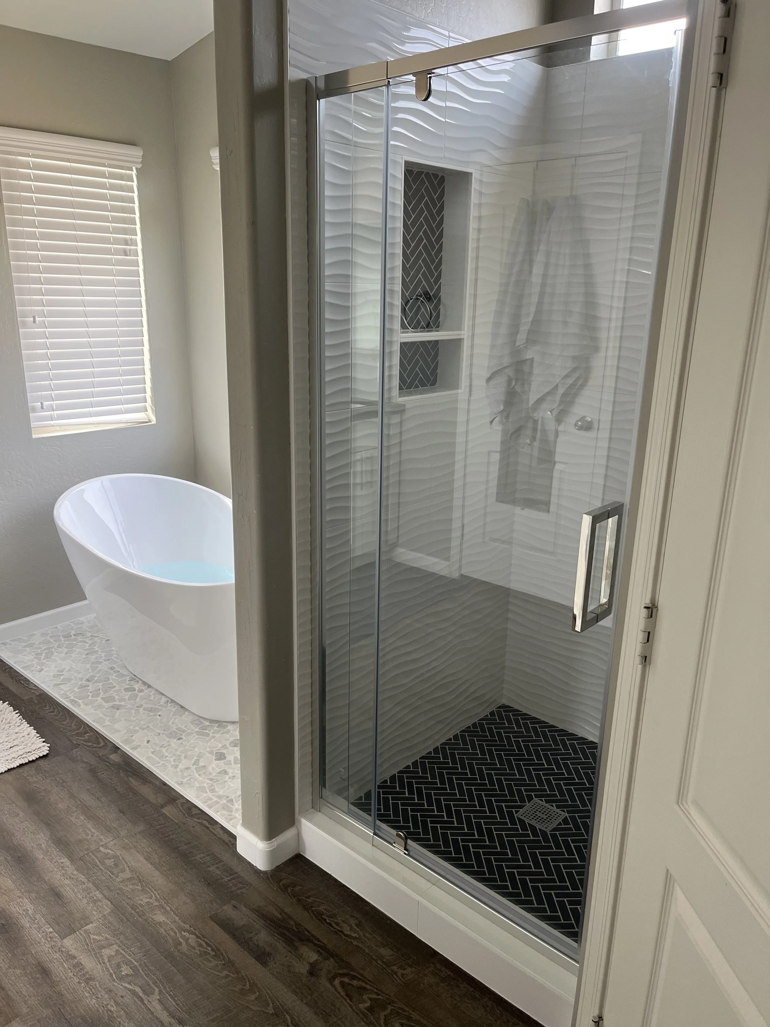 A bathroom with a glass shower enclosure with black herringbone tiles, a built-in wall niche with dark herringbone tile, a white bathtub near a window with white blinds, and wood-style flooring.