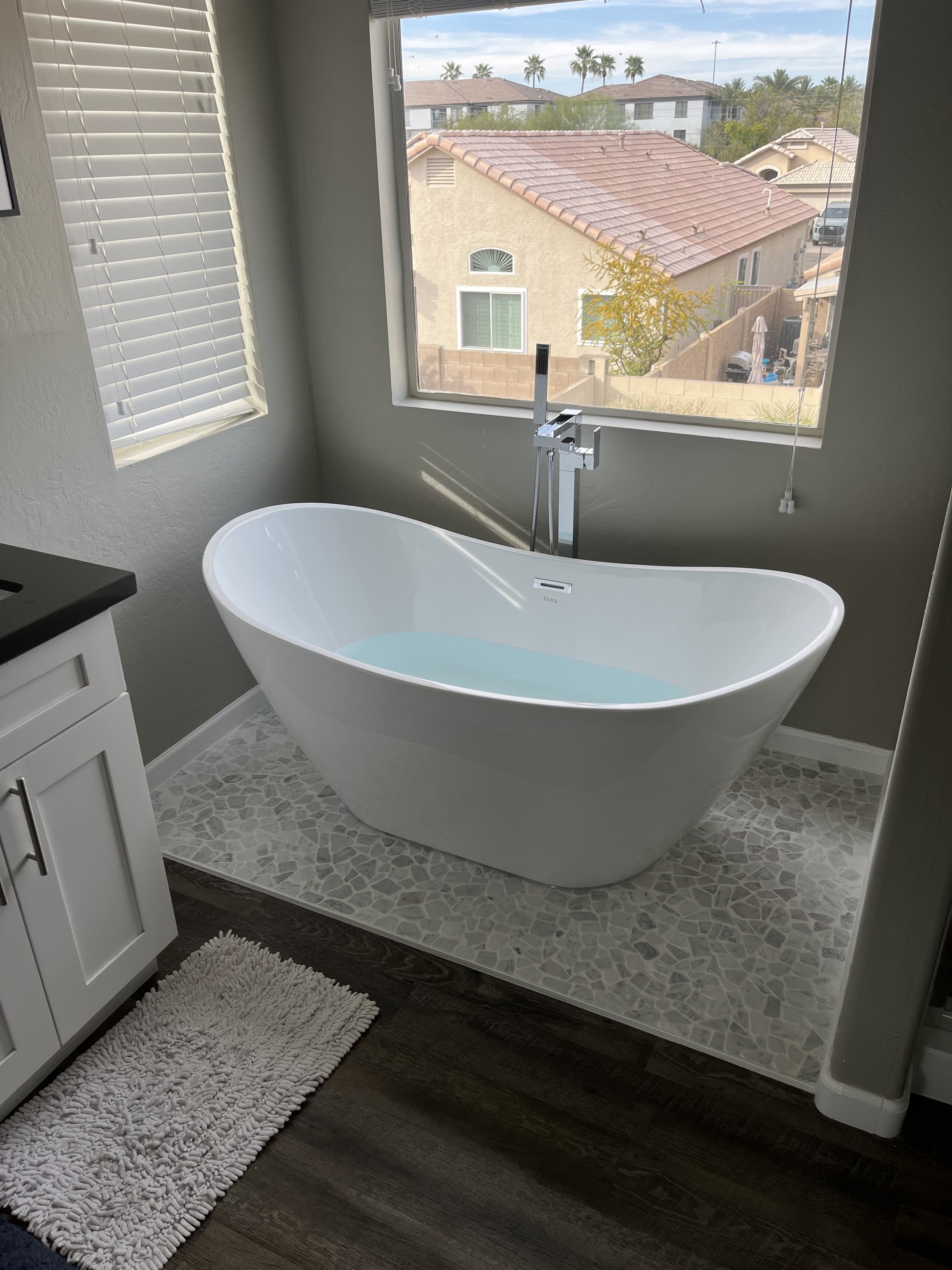 Modern bathroom with a white freestanding bathtub filled with water, a window with white blinds, dark wood flooring, a white cabinet with black countertop, a textured white rug, and outside view of neighboring houses and palm trees.