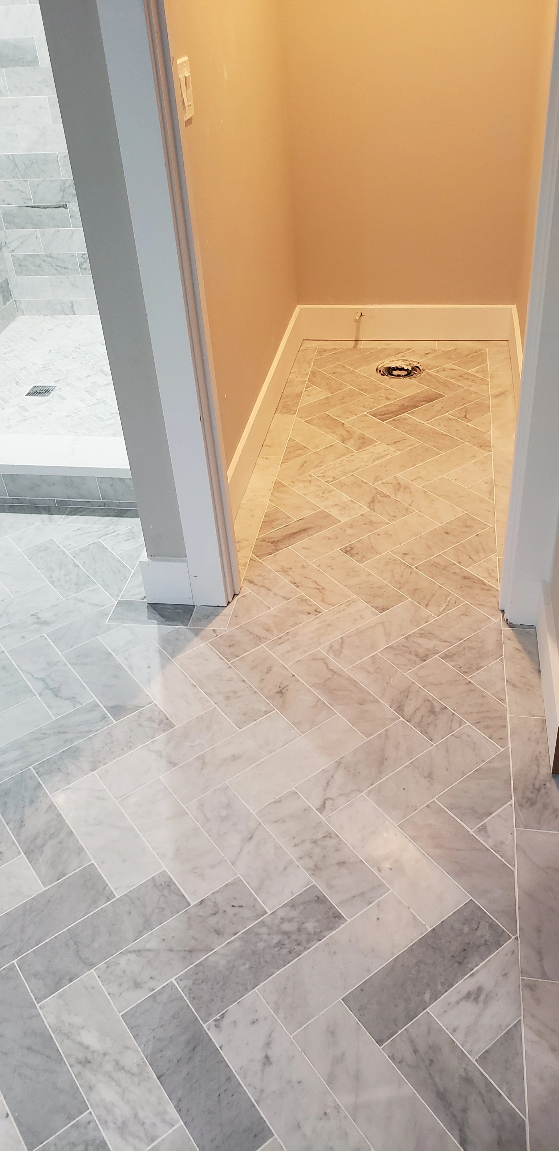 A room with marble tile flooring, a drain in the middle, and beige walls, adjacent to a shower area with similar marble tiles.