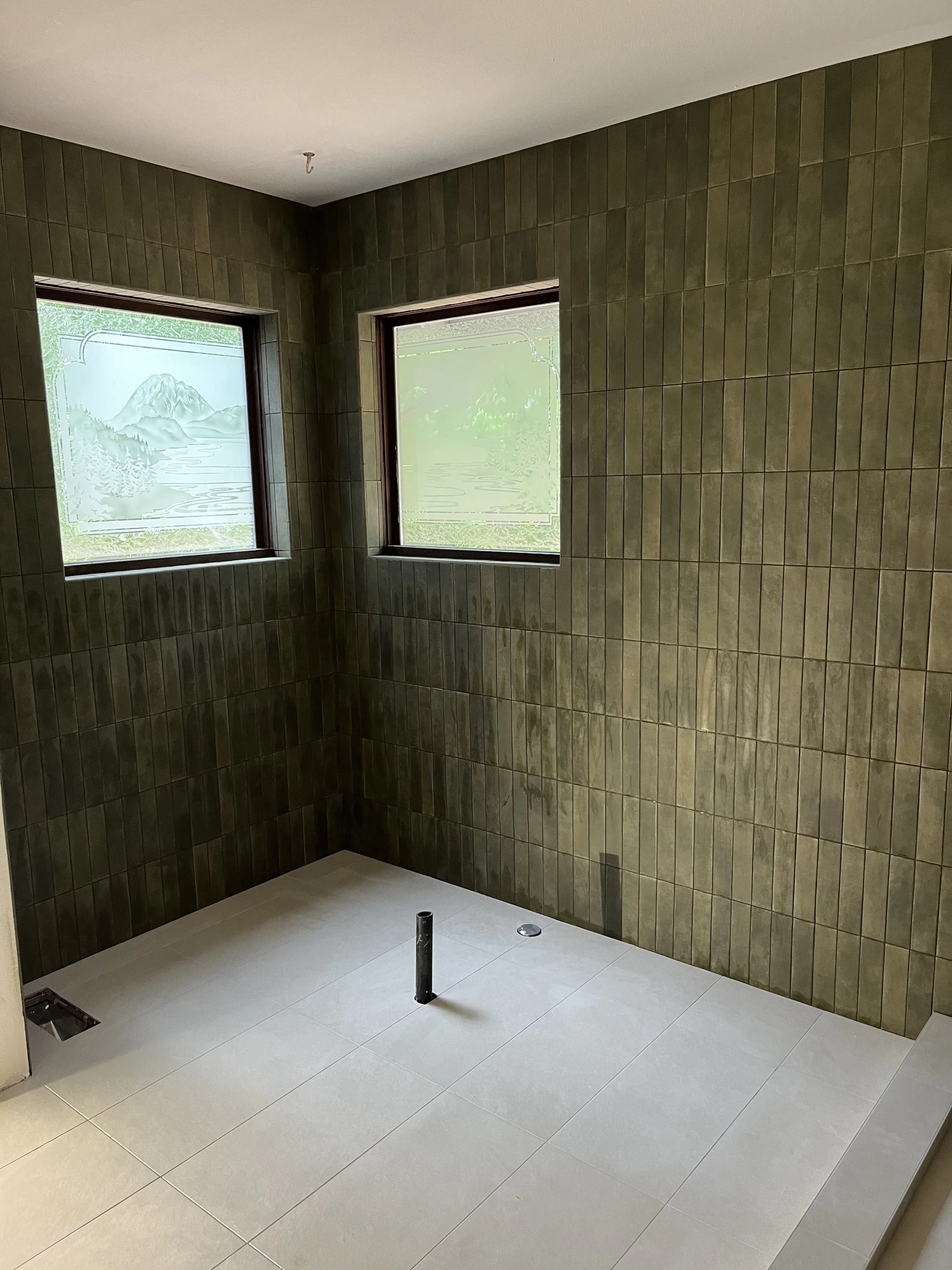 Empty room with beige floor tiles, brown tiled walls, two windows with mountain and lake landscape stickers, and plumbing pipes protruding from the floor.