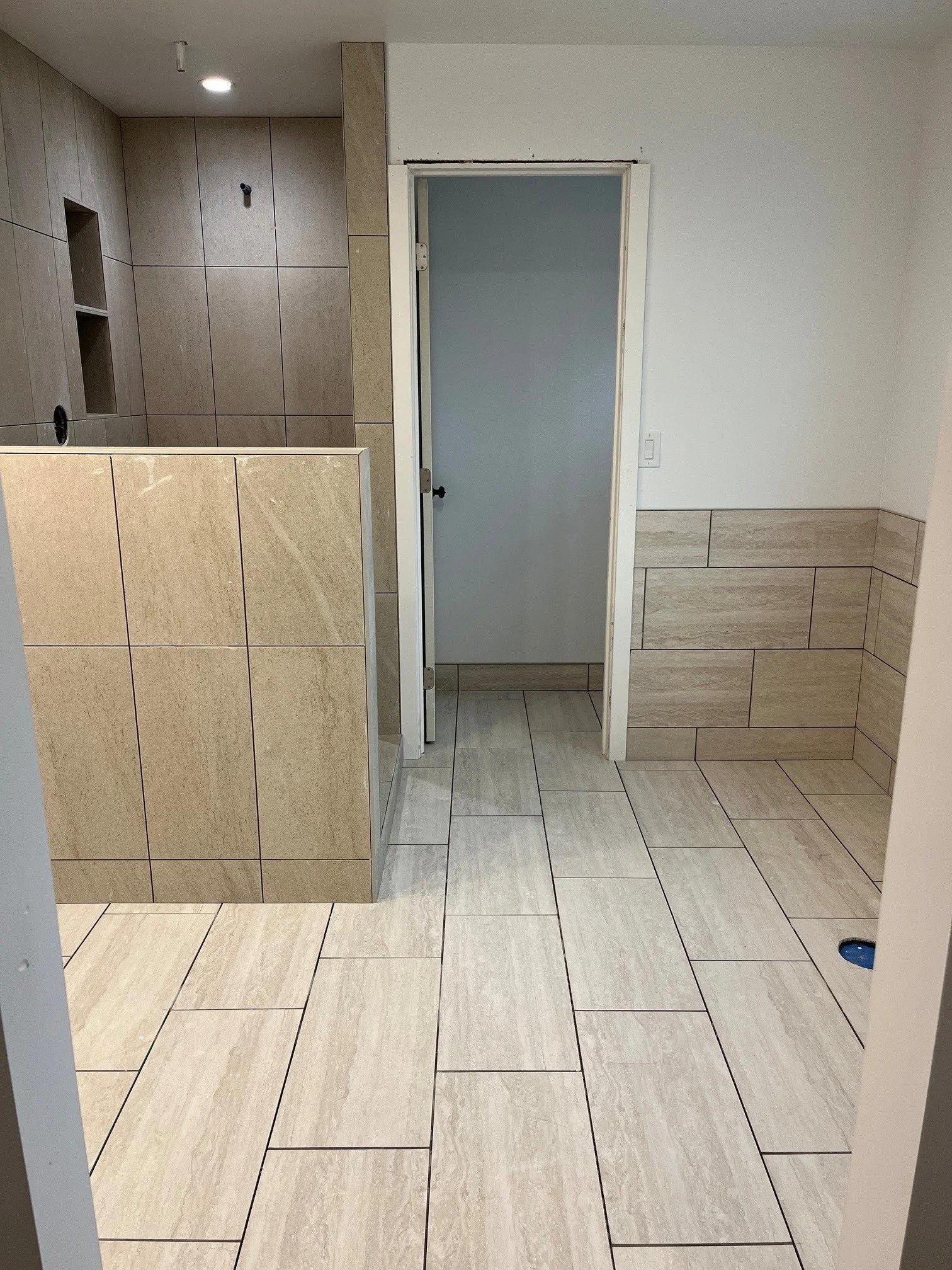 Public restroom stall bathroom with tiled floor and walls, white door with black doorknob, and an accessible sink area.