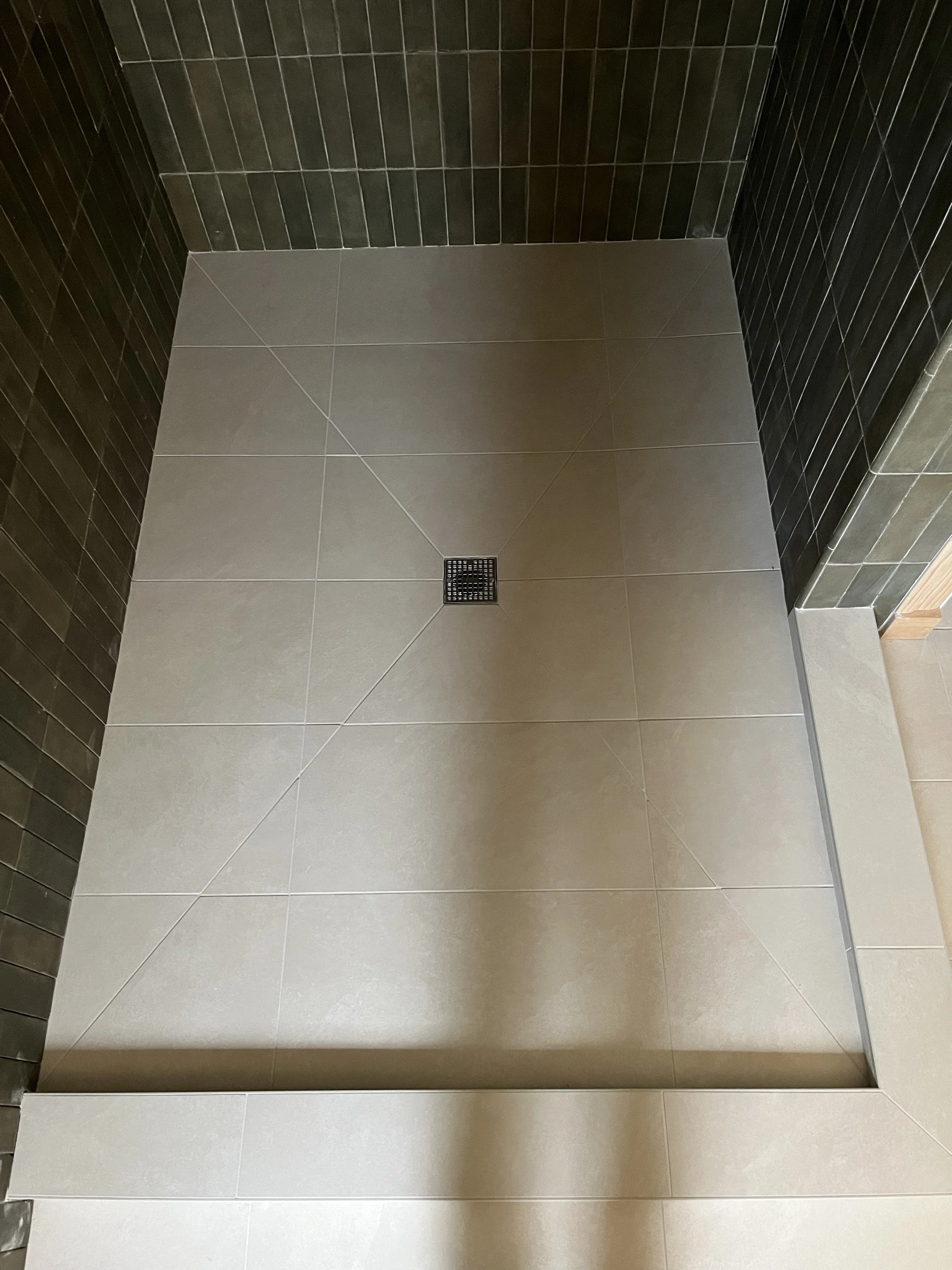 The image shows a modern, clean shower area with light gray floor tiles and dark gray wall tiles. There is a metal drain at the center of the floor. The shower space appears to be at a corner with both side walls visible.
