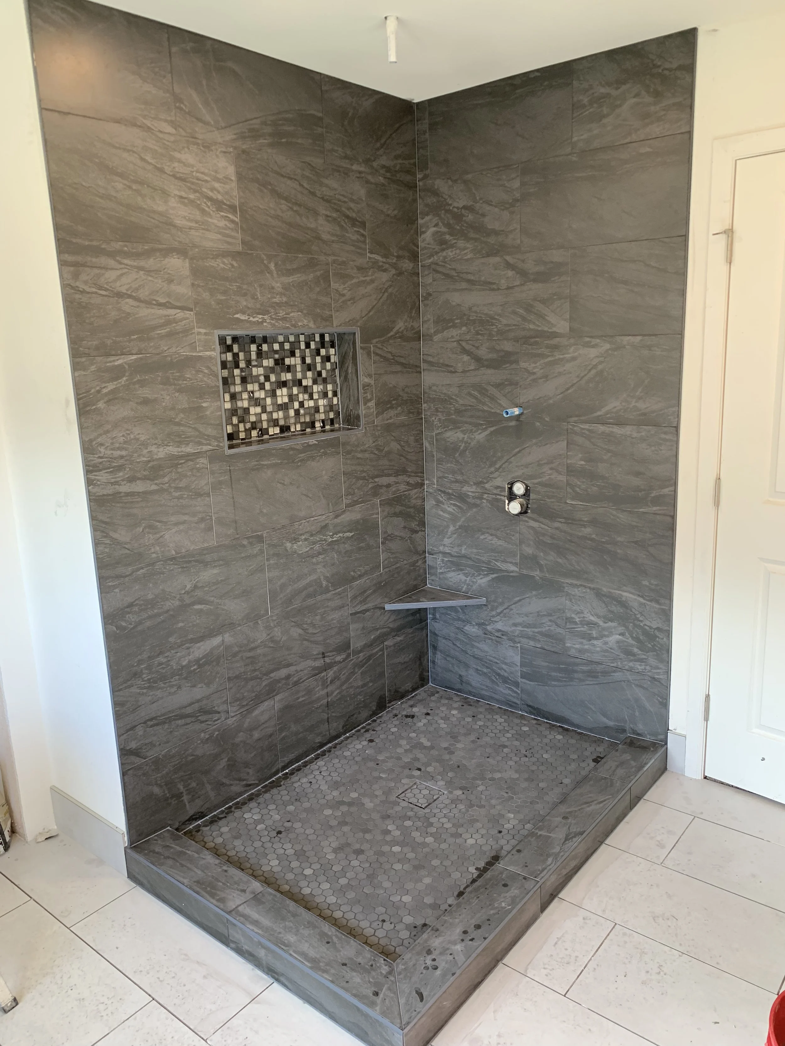 Bathroom shower with gray marble tiles on the walls, a small recessed shelf with mosaic tiles, and a hexagonal tiled shower floor. The shower area is partially enclosed with a doorless design.