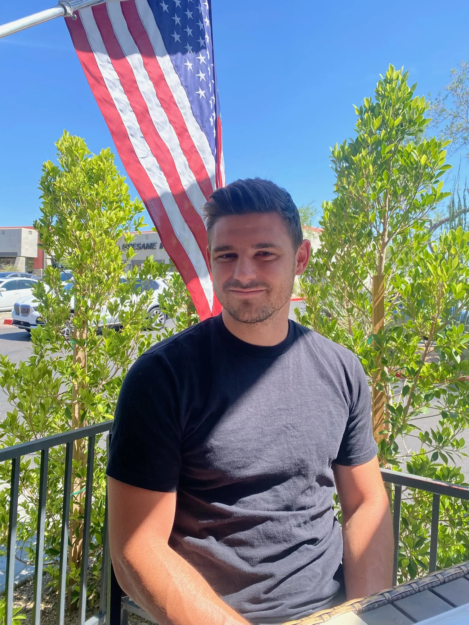 A young man with short dark hair and a stubble beard is sitting outdoors at a table, wearing a black and gray t-shirt. An American flag is waving on a pole in the background, along with green bushes, a clear blue sky, a parking lot with white cars, and a building with the