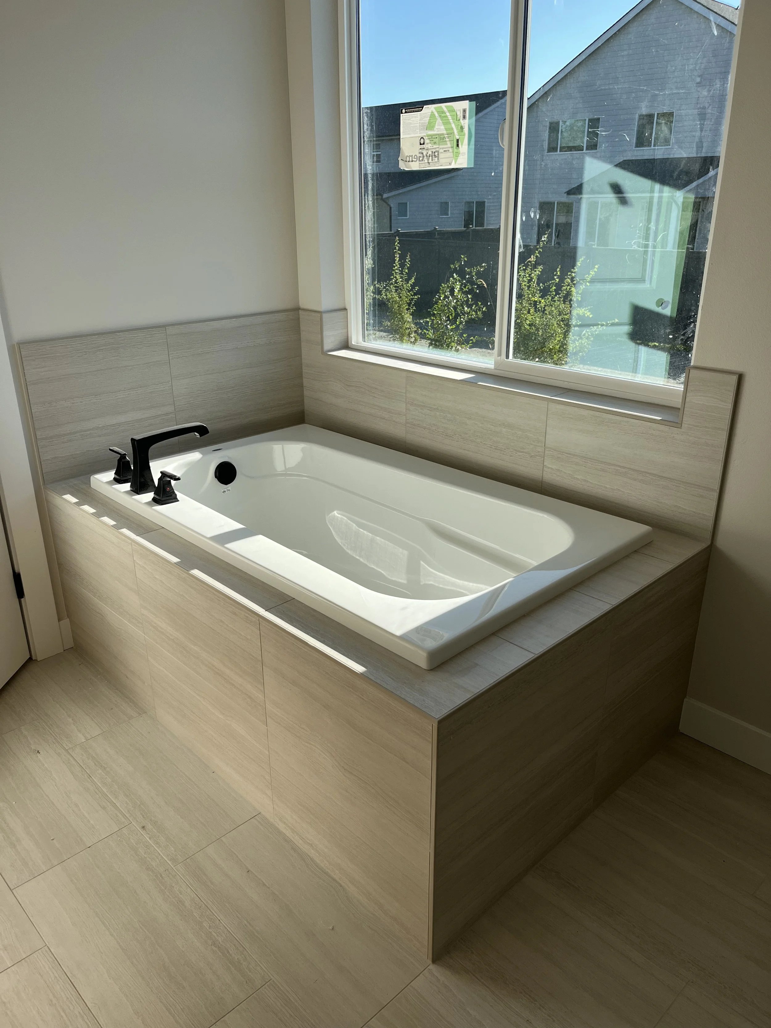 A modern bathroom with a built-in rectangular bathtub, black faucet, and a large window showing neighboring houses and greenery.