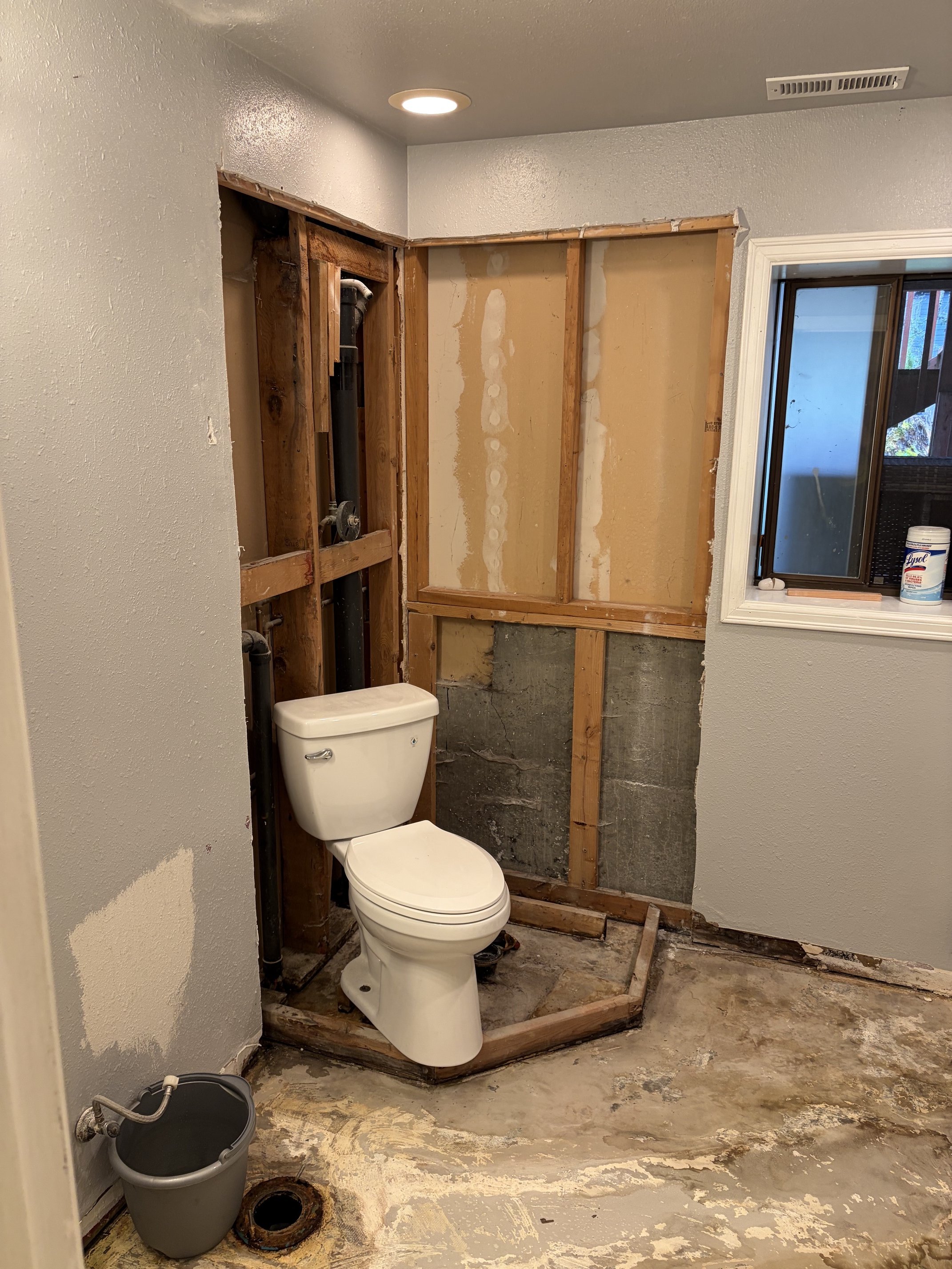 Bathroom under renovation with exposed wall framing, partial drywall, a toilet, a bucket, and construction debris.