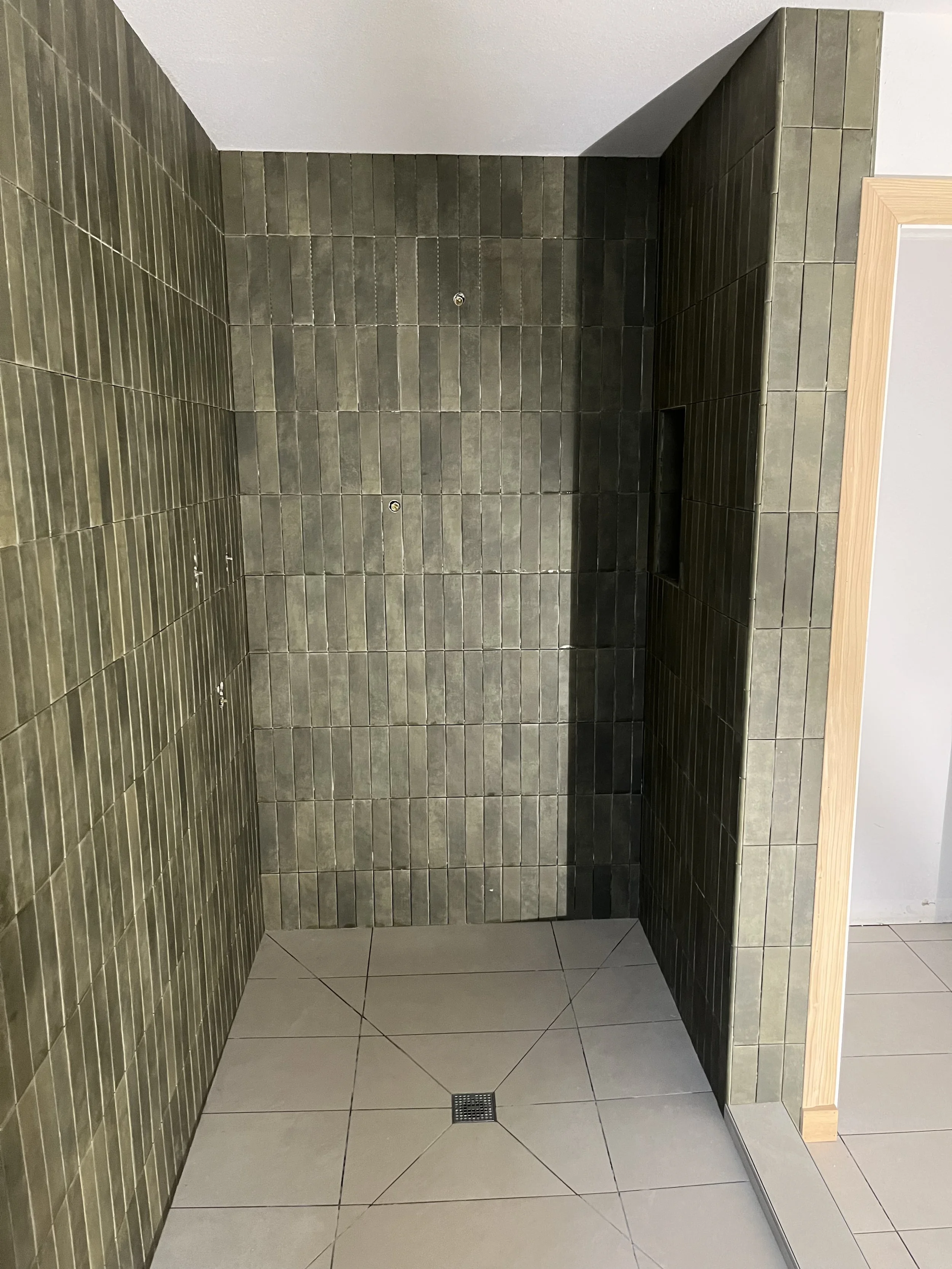 A shower stall with green tiled walls, a drain on the white tiled floor, and a small built-in shelf in the wall.