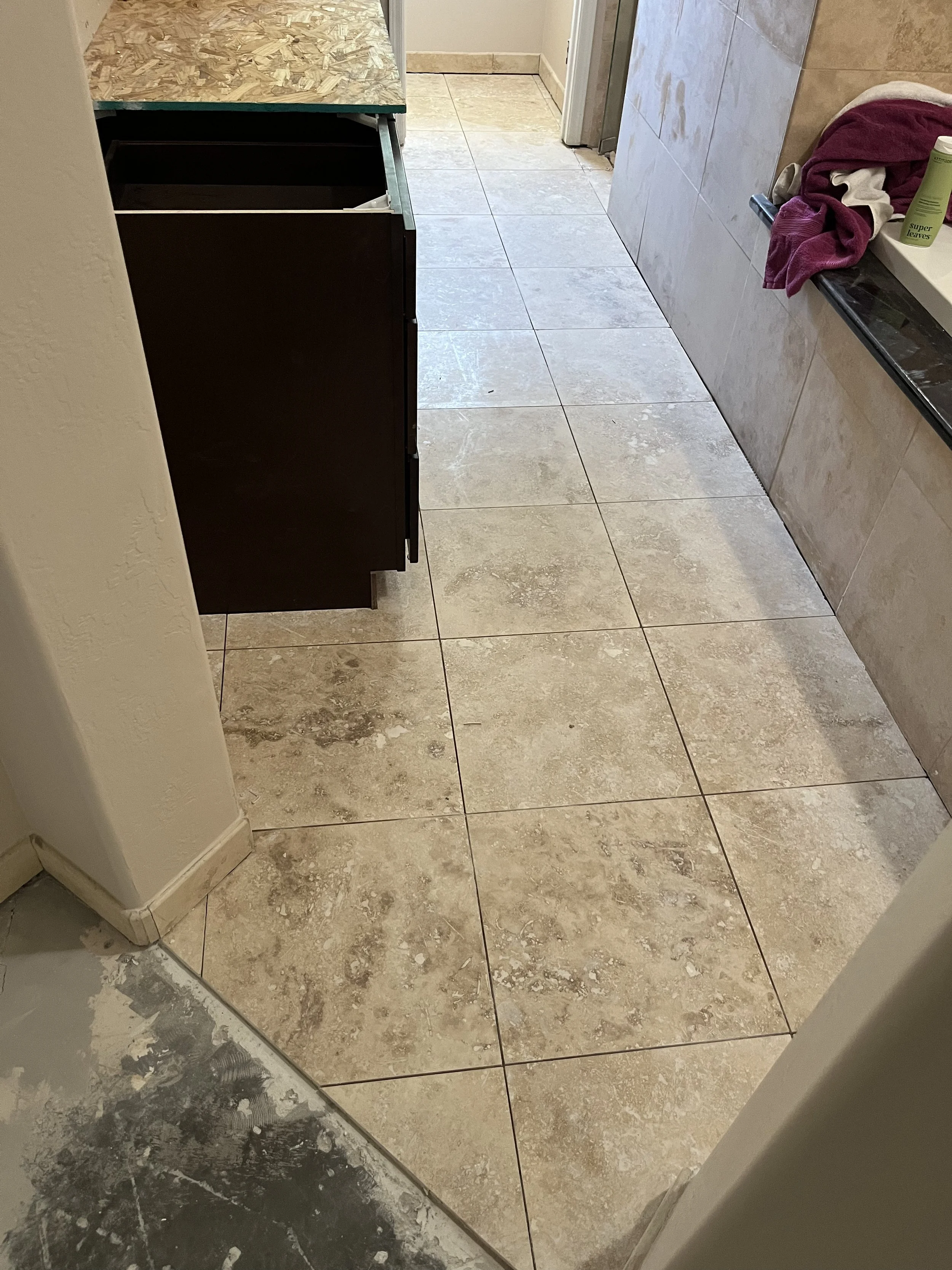 Bathroom with beige tiled flooring, a black cabinet, a towel on the brown marble countertop, and some construction materials on the floor.