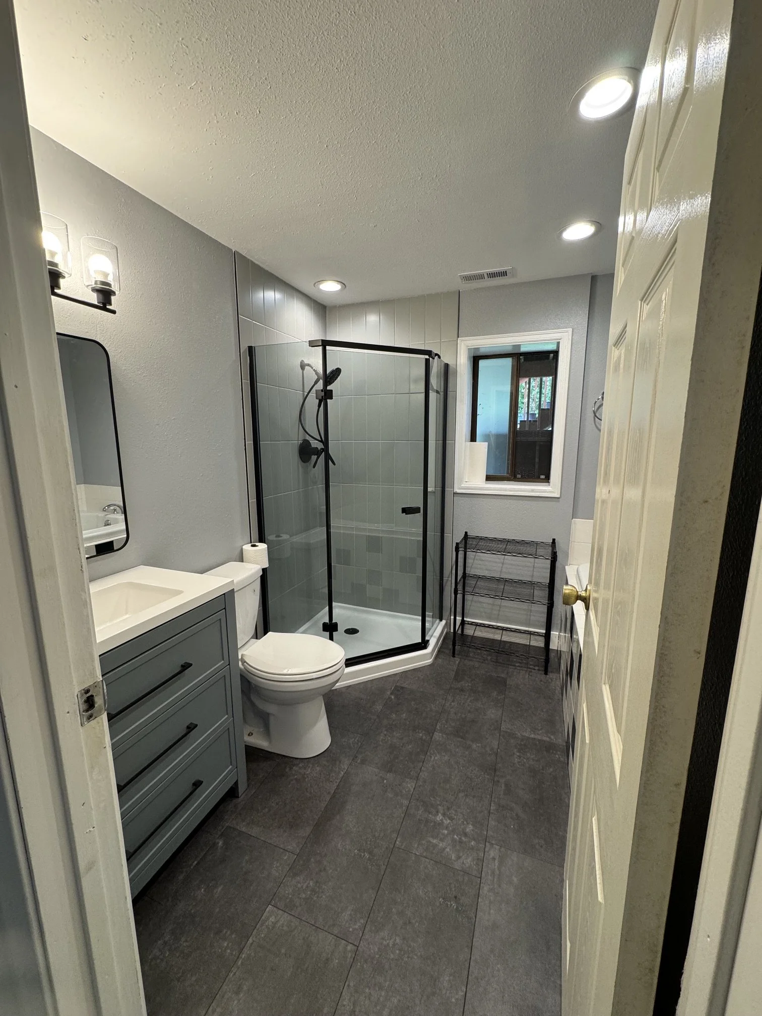 Bathroom with a shower stall with glass doors, a white toilet, a small vanity with a mirror and faucet, a window with trees outside, a black metal rack, gray walls, and dark gray tiled floor.