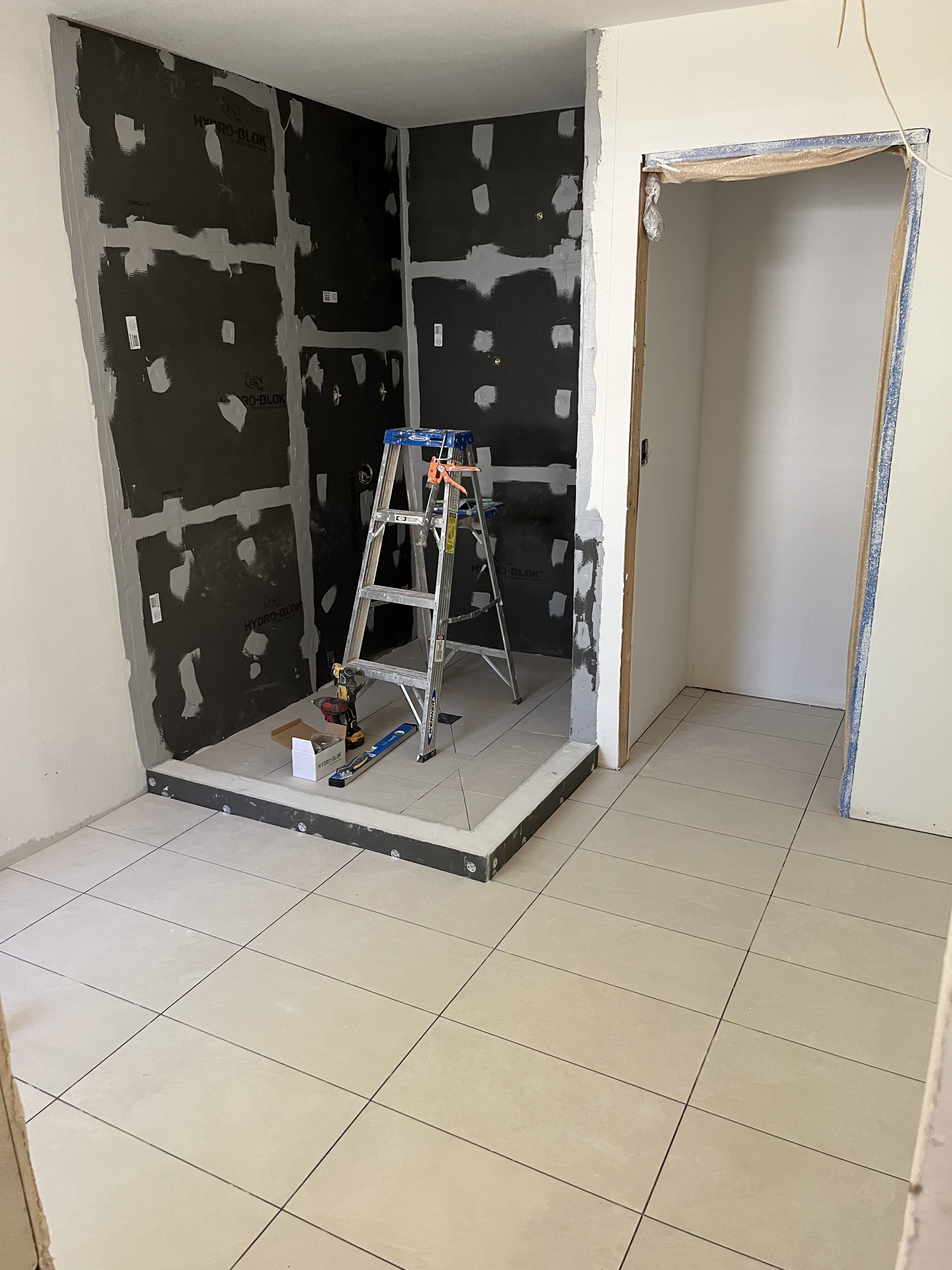 Interior of a space under renovation, showing a partially constructed shower or bathtub area with a black waterproofing membrane, construction tools, and a step ladder inside. The surrounding walls are being prepared for tiling or finishing.