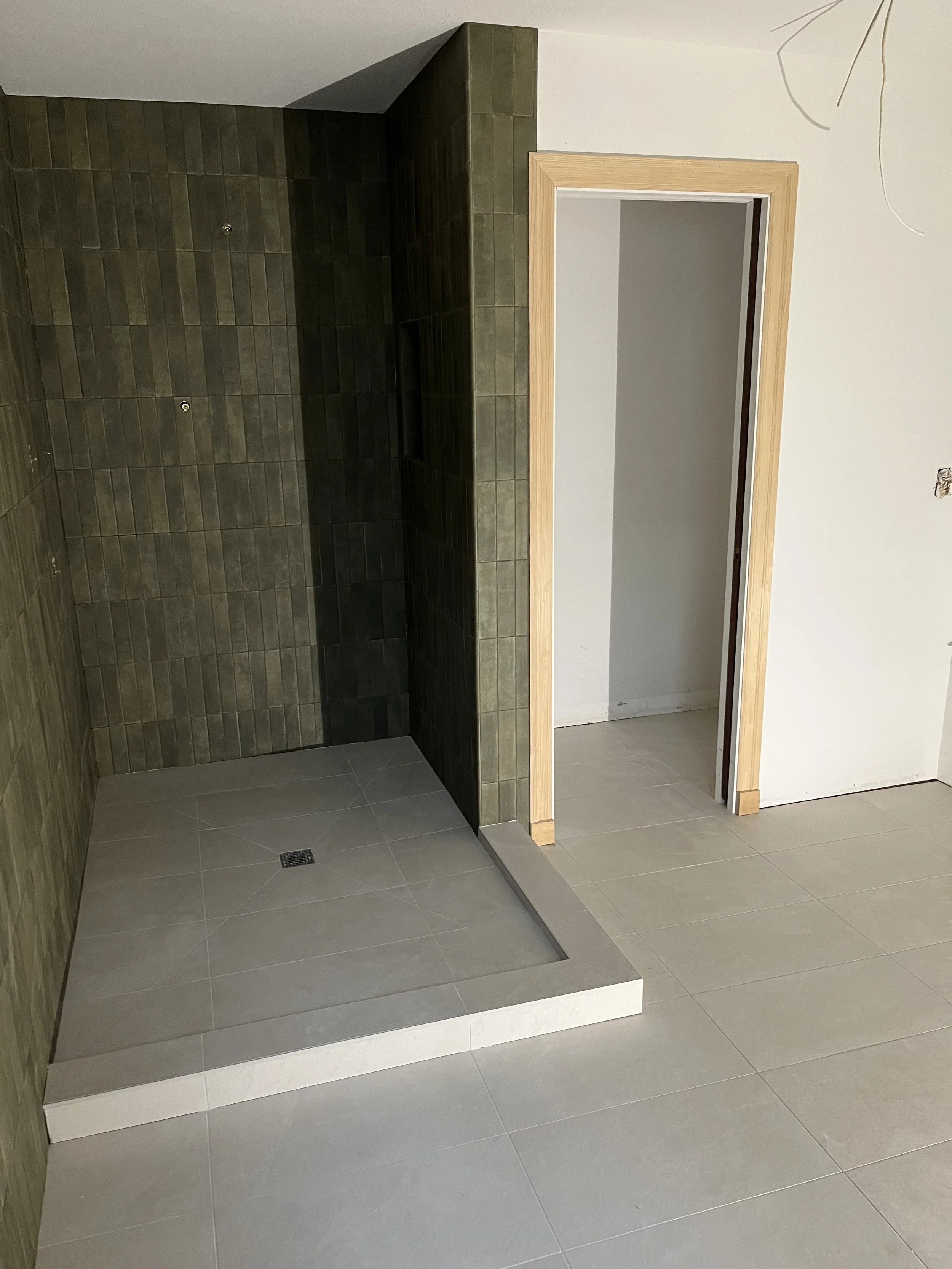 A partially constructed bathroom shower with green tiles on the walls, a floor drain, and a wooden door frame without a door installed yet.