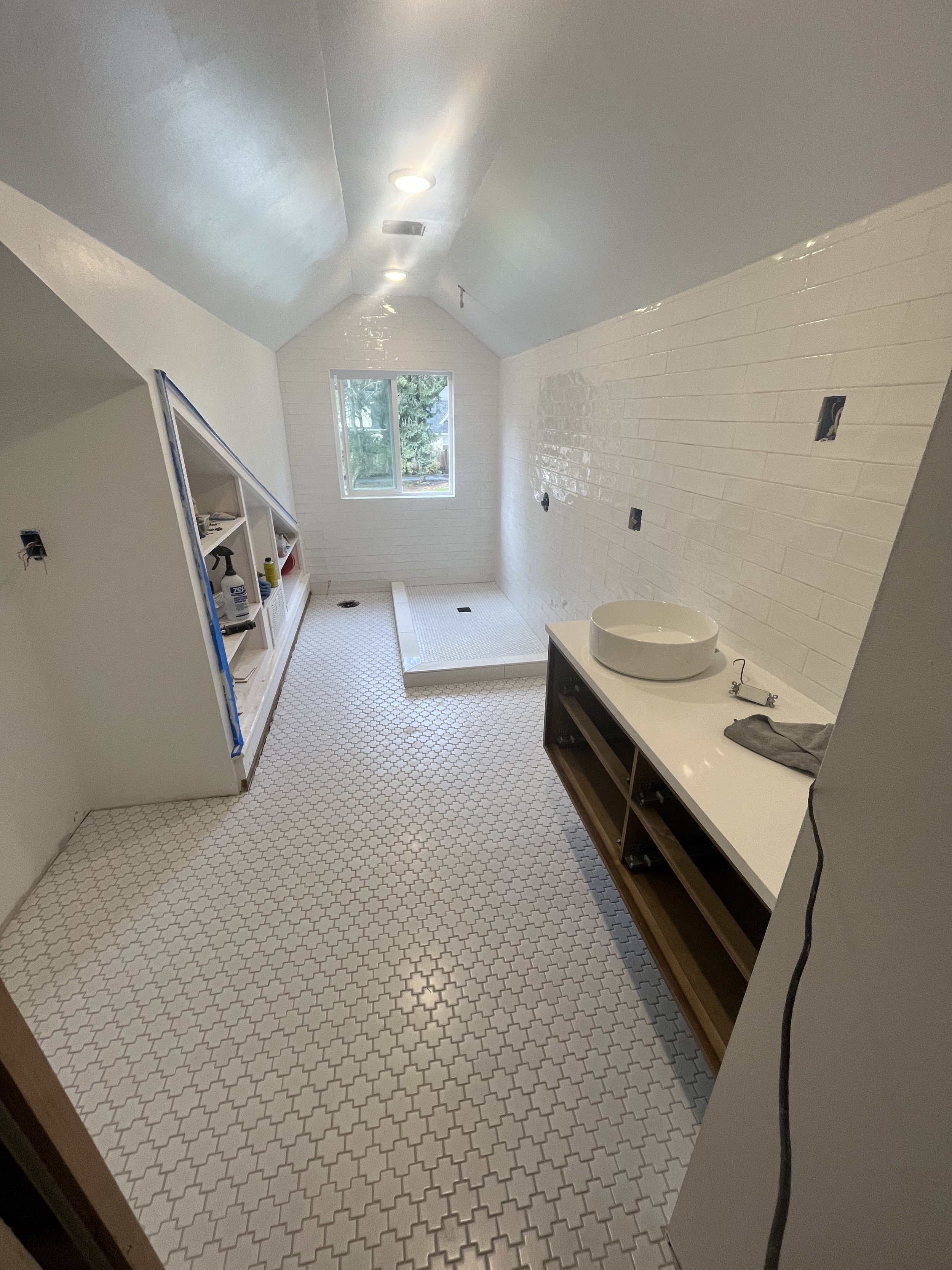 A bathroom under construction with white tiled walls, a window showing trees outside, and a patterned floor. There is an open shelf with supplies, a shower area, and a vanity with a round vessel sink.