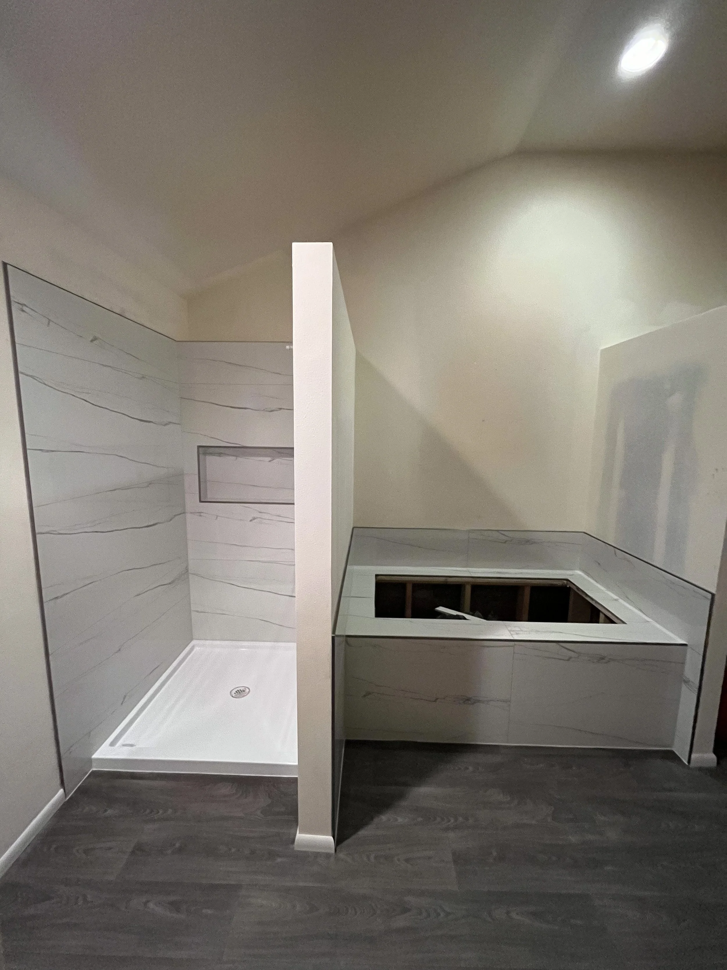 A bathroom with a walk-in shower featuring white marble walls and gray veining, a drain on the floor, and a partial view of a vanity with a large mirror framed in gray. The room has wood-like dark flooring and a light-colored ceiling with ceiling lig