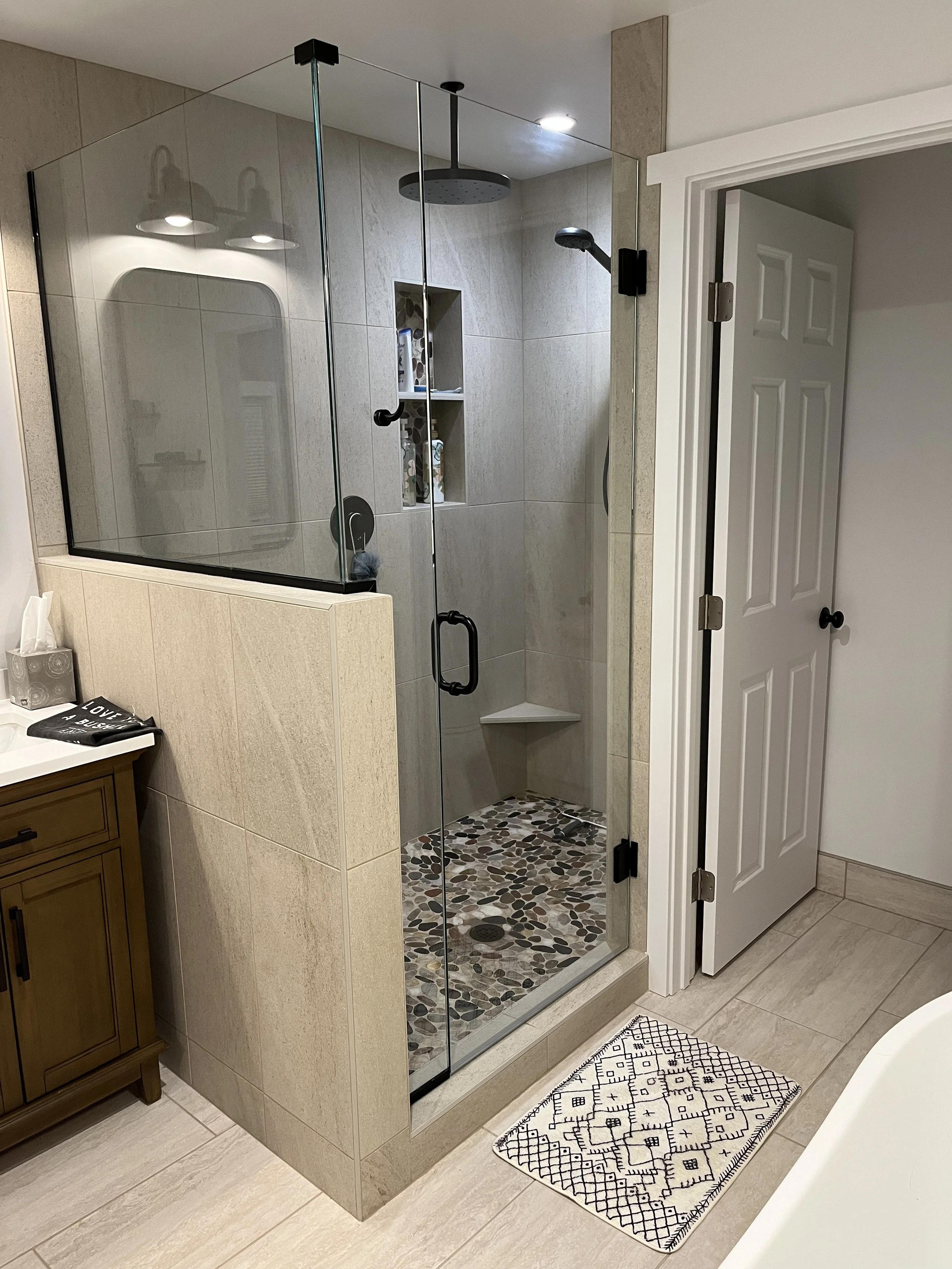 A modern glass shower stall with beige tiles and pebble-style floor tiles, located in a bathroom next to a white door. The shower features a large rain shower head and a hand-held shower spray, with built-in shelves holding toiletries. A decorative b
