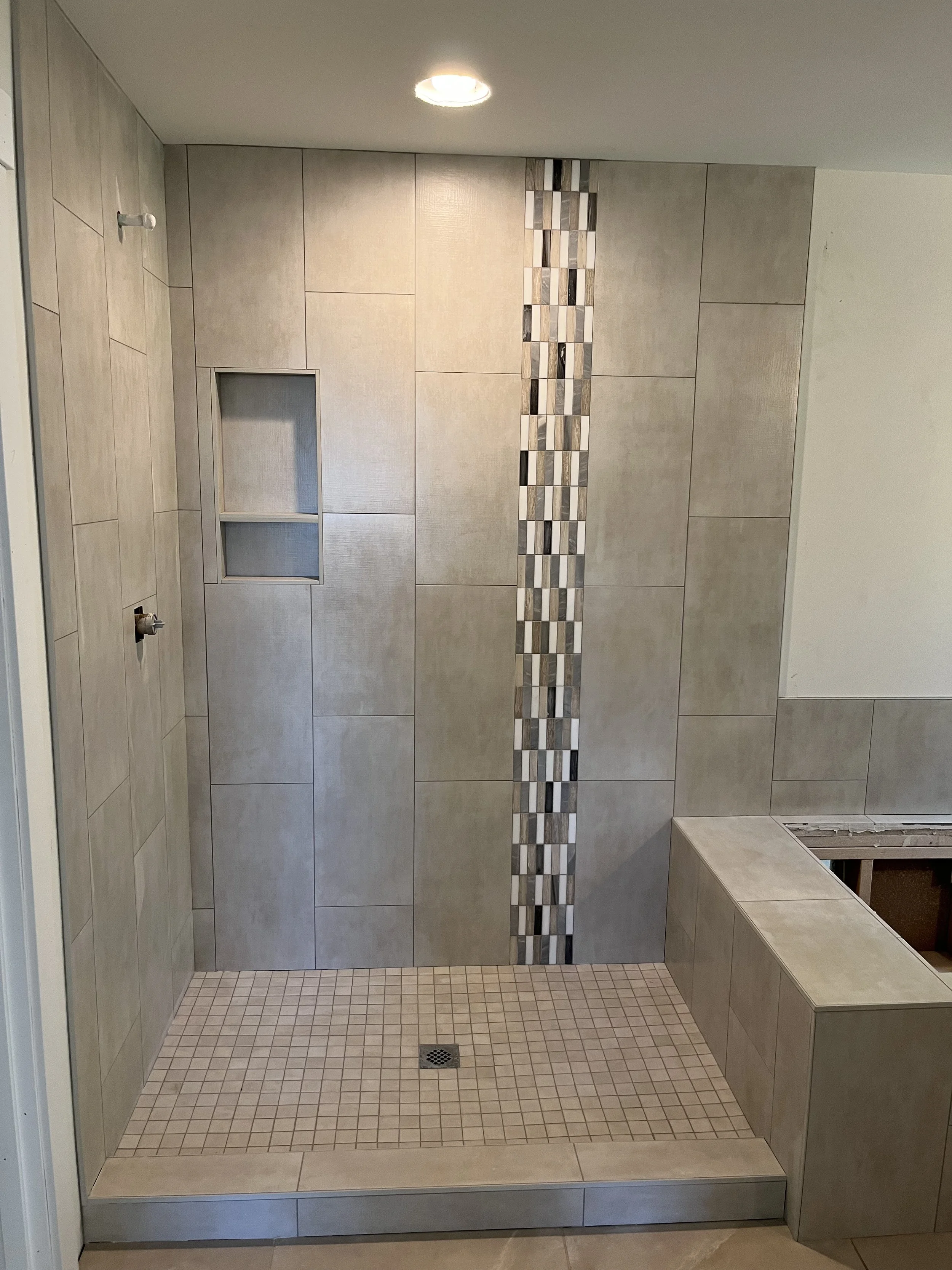 A tiled shower area with a built-in bench on the right side, a niche for toiletries on the left wall, and a vertical strip of decorative mosaic tiles running down the center of the back wall. The floor has small square tiles with a drain in the middl