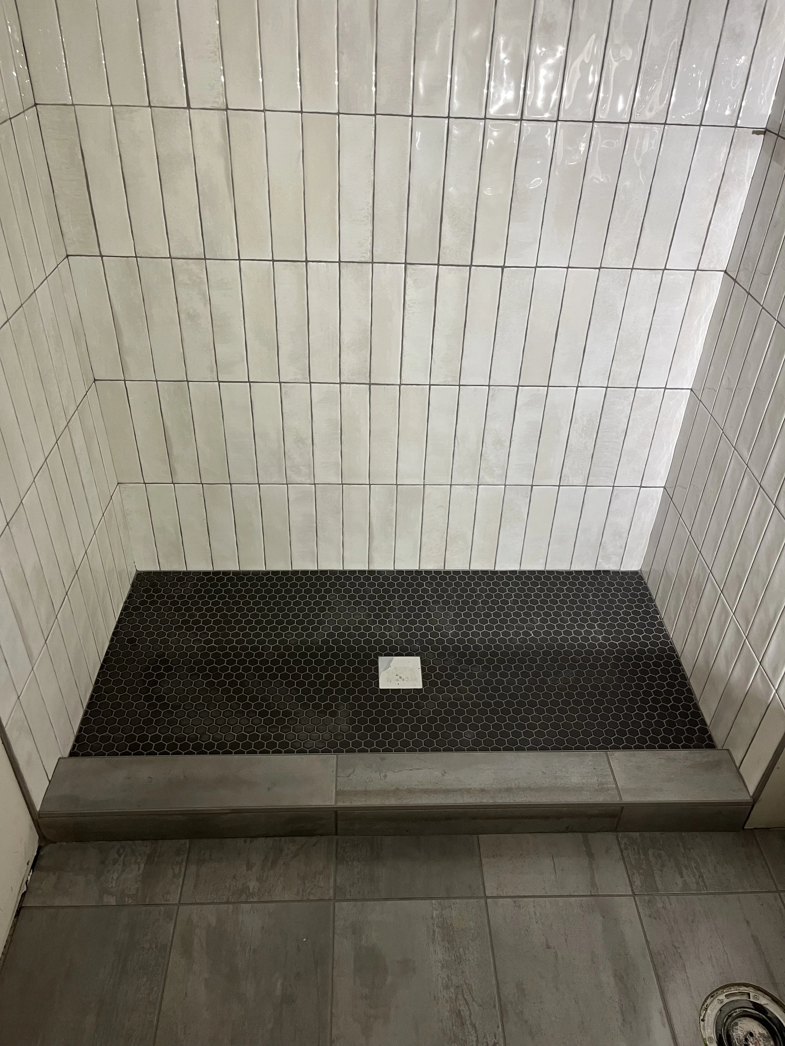 The image shows a shower area with white rectangular tiles on the wall, small black hexagonal tiles on the floor, and a drain on the floor near the center.