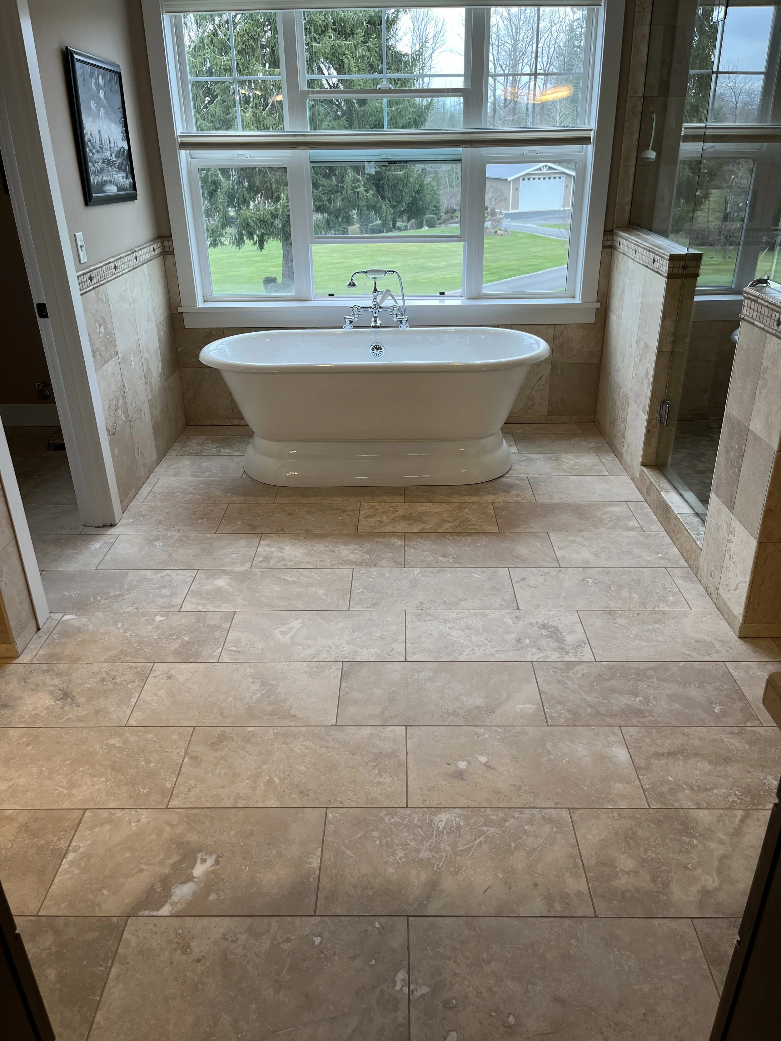 A bathroom with a white bathtub placed in front of large windows showing a green lawn and trees outside. The bathroom features beige tile flooring and marble tile walls, with a glass shower enclosure on the right side.