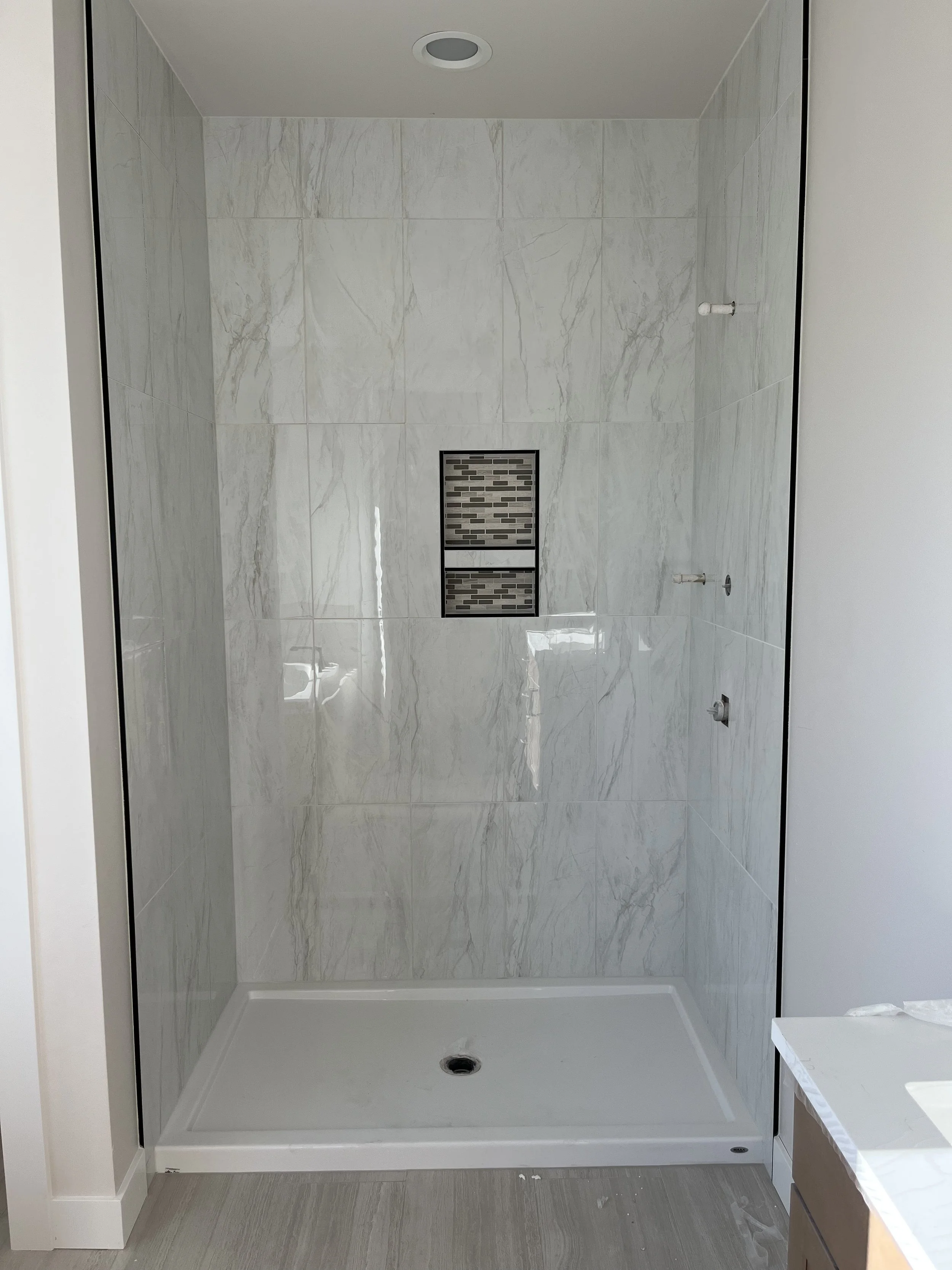 Empty shower area with marble wall tiles, a drain in the floor, and a built-in niche with mosaic tile pattern. The shower has no fixtures installed yet.
